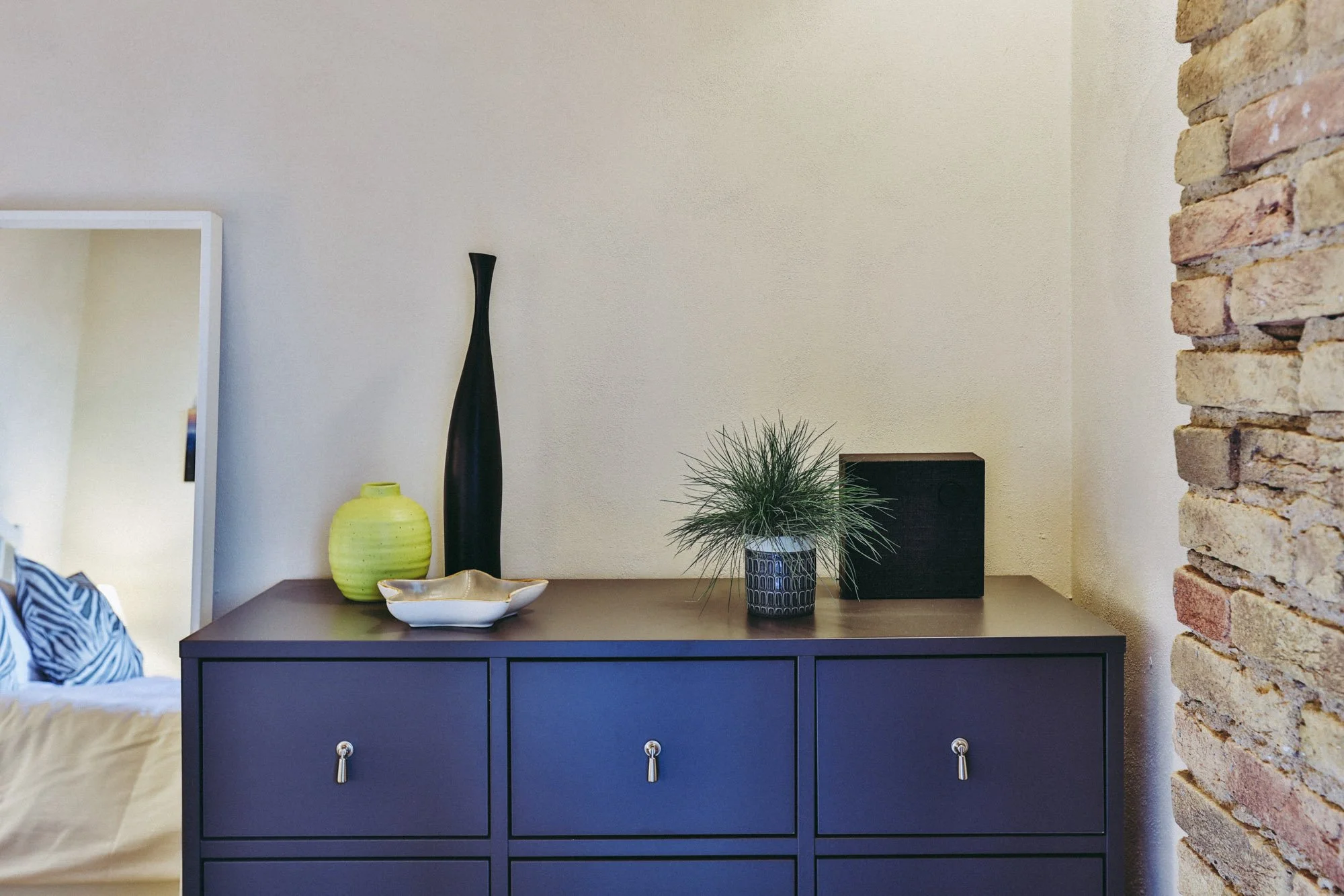A gray cabinet with two drawers and silver handles sits against a beige wall decorated with vases and plants, with a brick wall on the right side.