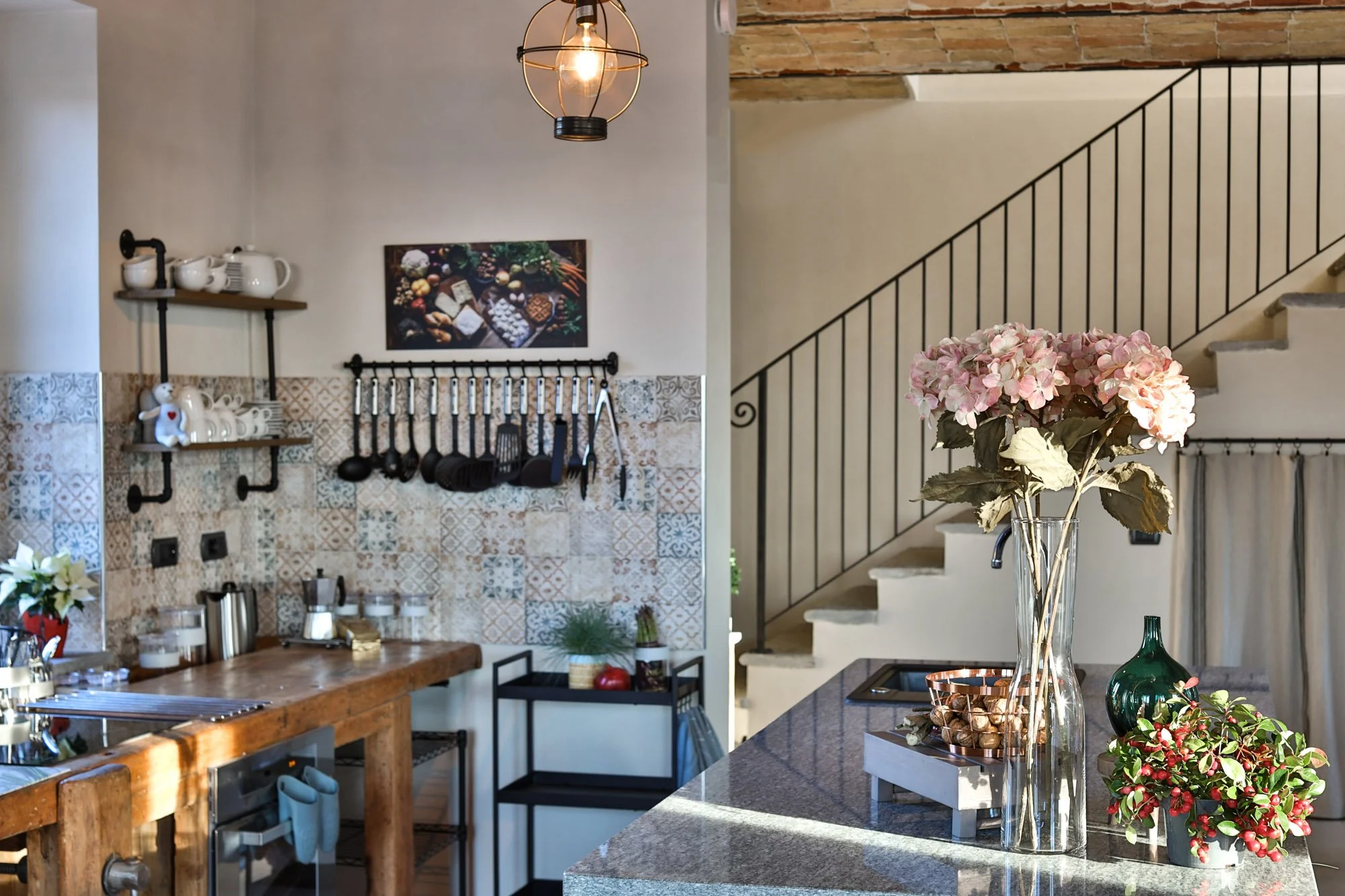Modern kitchen with granite countertops, a glass vase with pink hydrangea flowers, and decorative plants, with a staircase and hanging light fixture in the background.