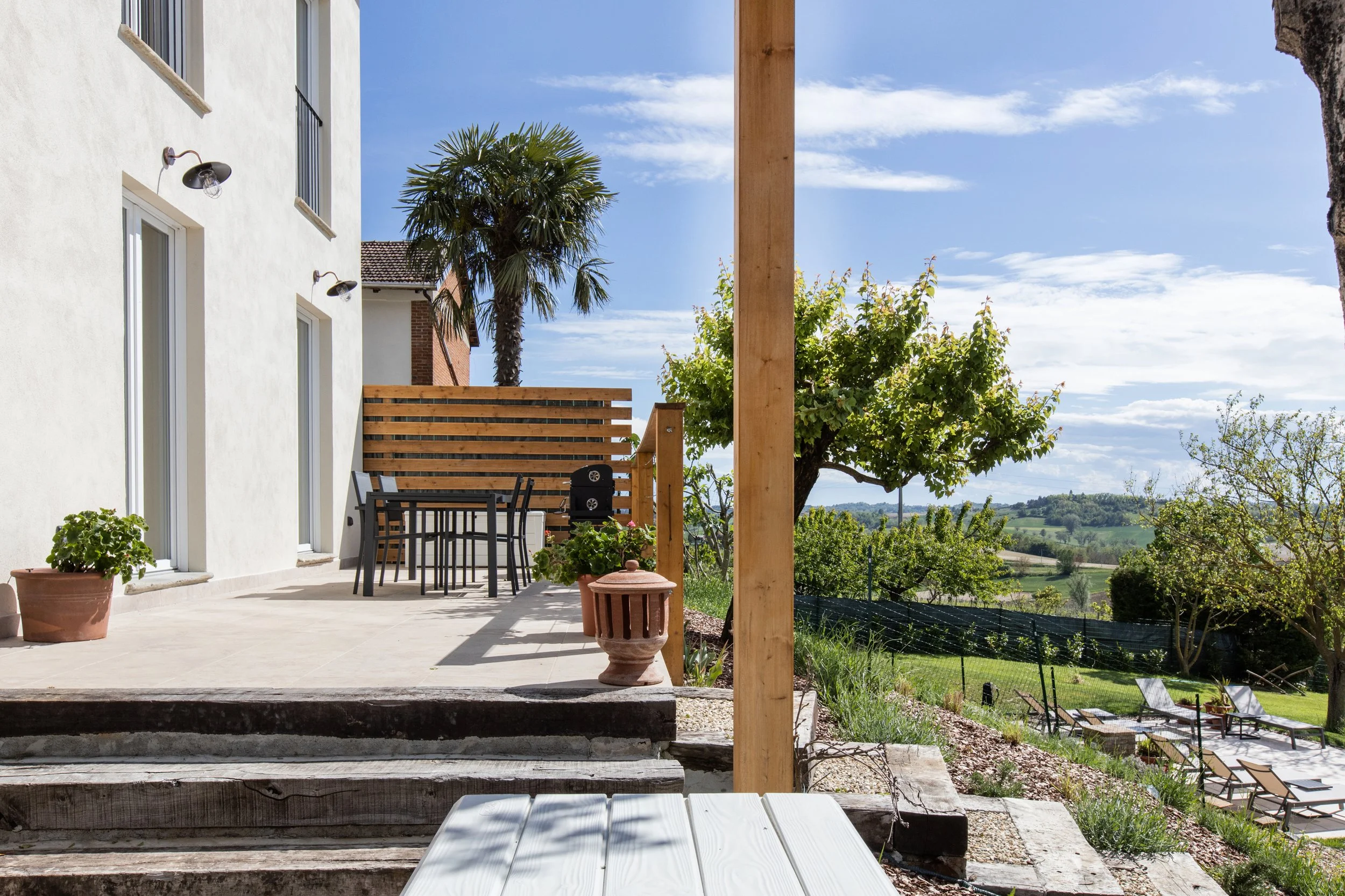 View of a backyard patio with outdoor table and chairs, potted plants, a palm tree, and scenic rolling hills in the background on a sunny day.