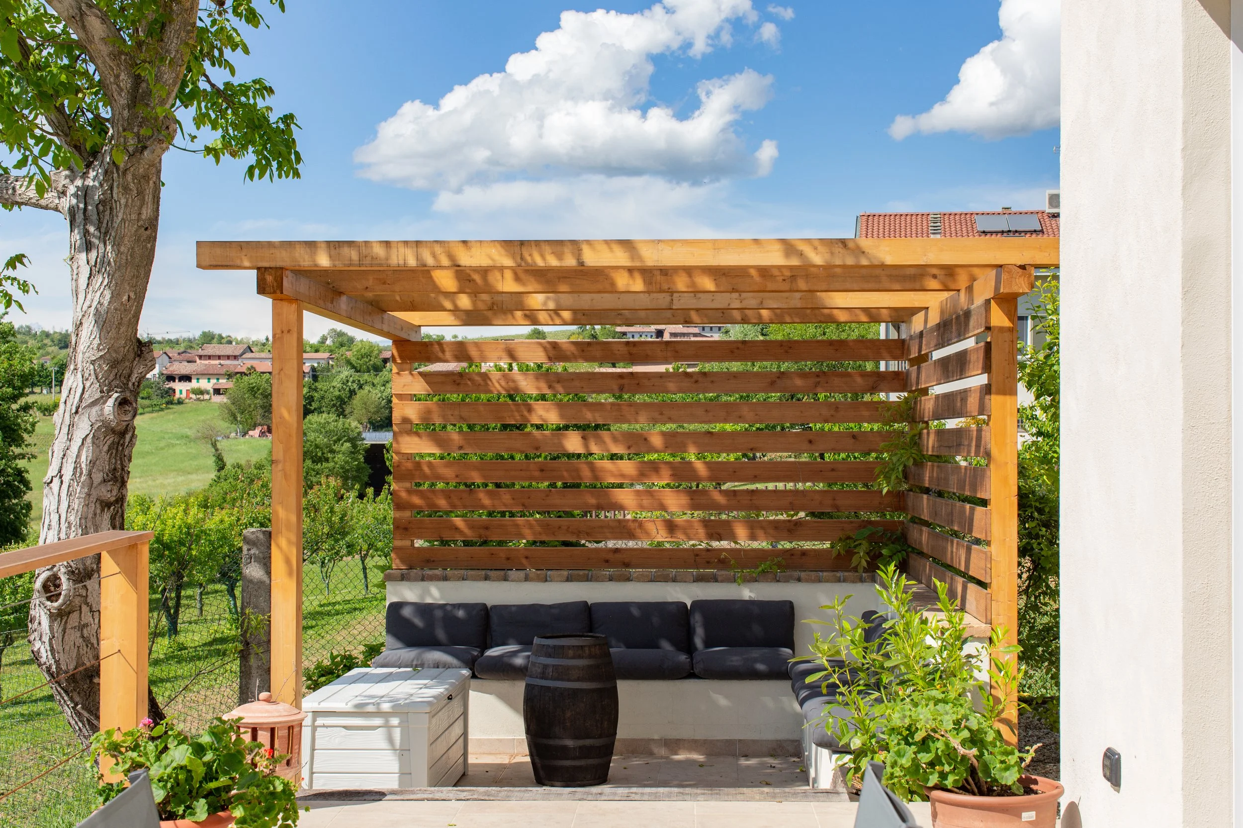 A small patio or seating area with a black sofa, a white cooler, and a black barrel-shaped table, surrounded by potted plants and greenery. A wooden pergola provides partial shade, and a large tree is visible on the left. The background features a vi