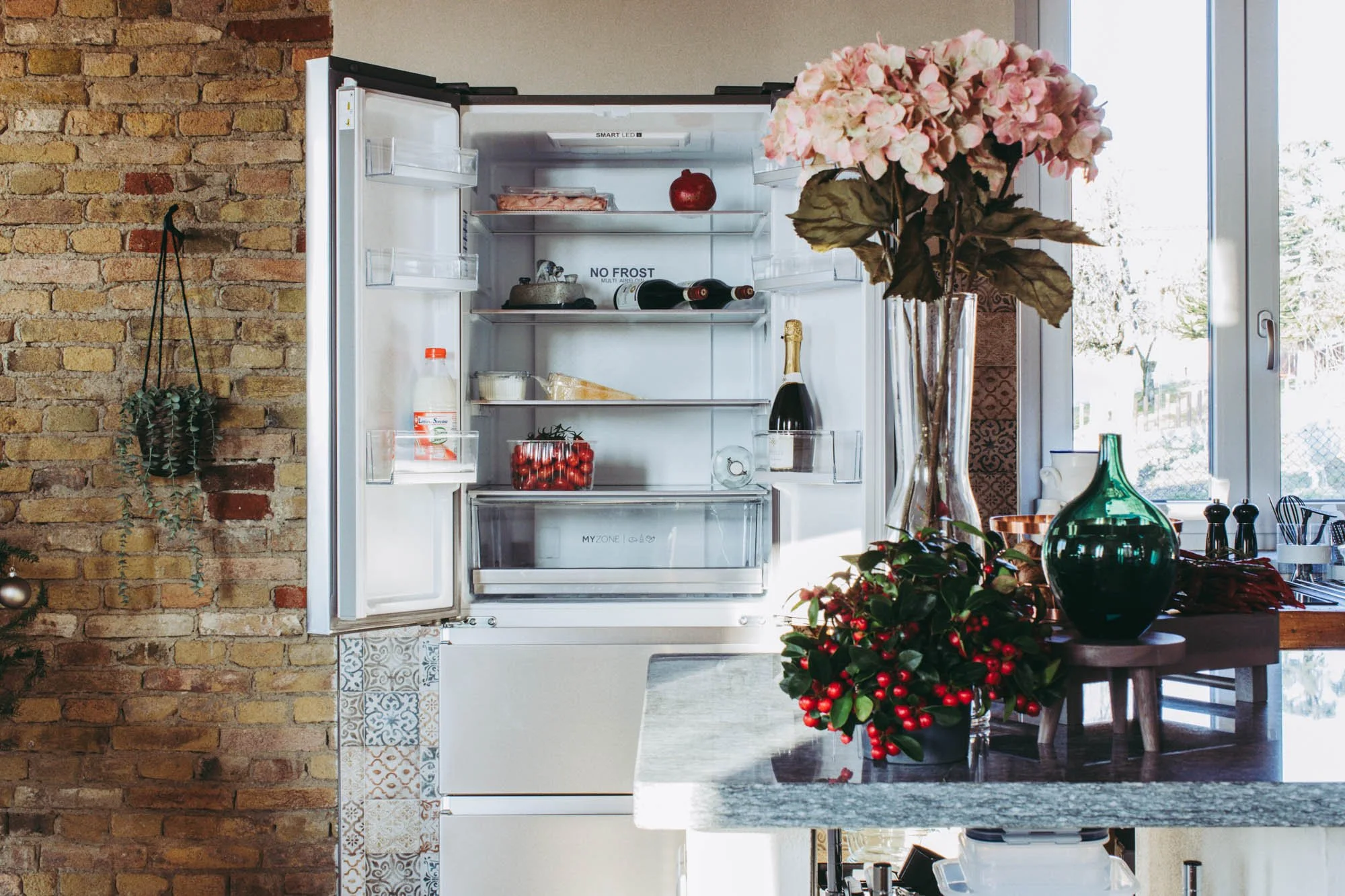 An open refrigerator in a kitchen, with a large vase of pink hydrangeas on the counter nearby, a brick wall to the left, and a sliding glass door to the right.