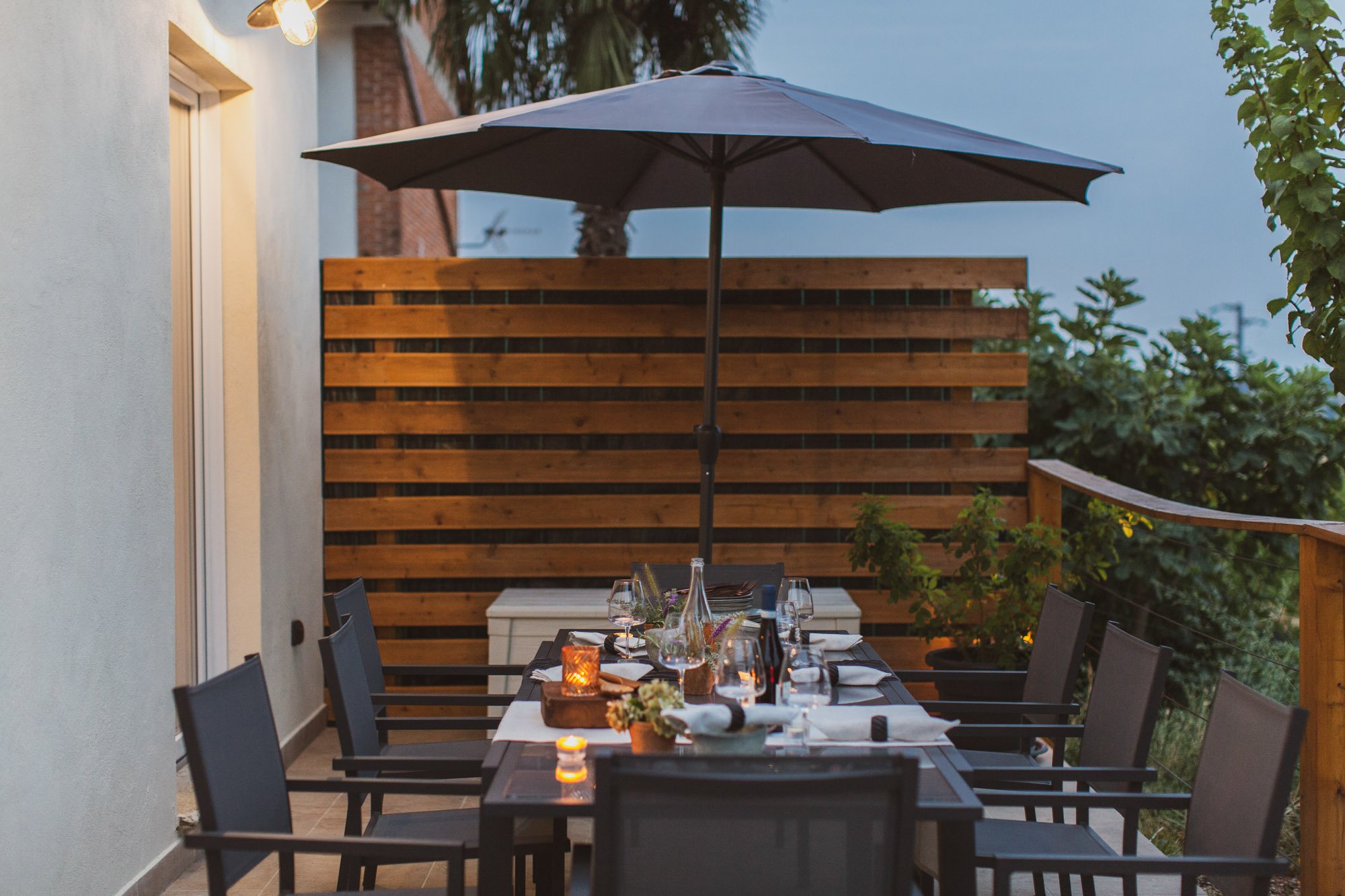 An outdoor dining area with a table set with wine glasses, a bottle of wine, candles, and flowers, under a large umbrella. There is a wooden privacy fence, greenery, and a white exterior wall with an outdoor light fixture.