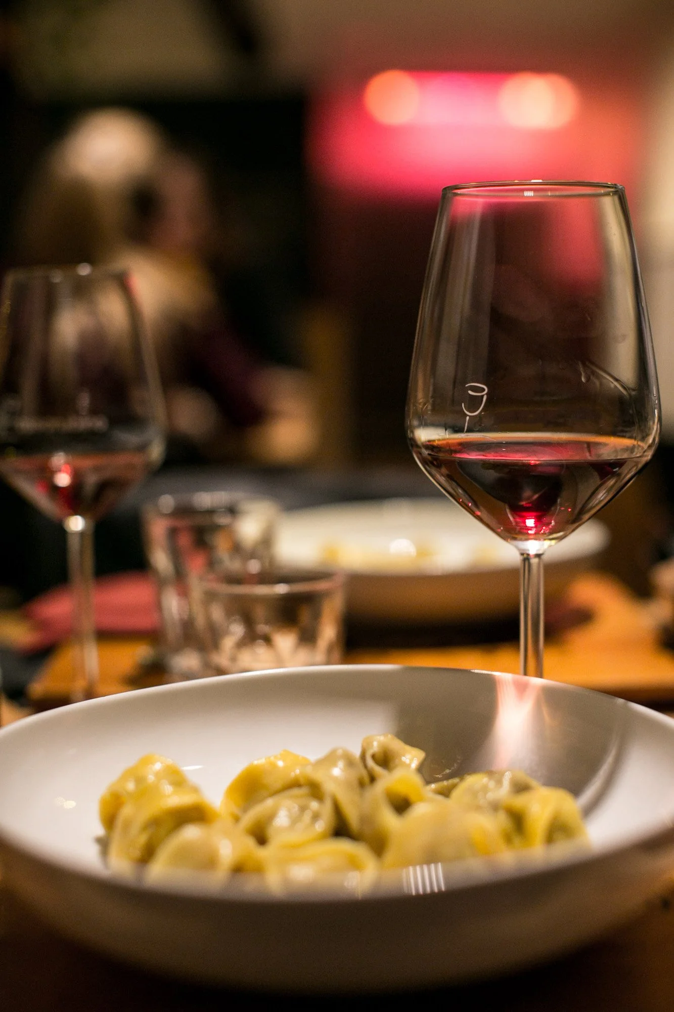 A glass of red wine on a table in focus, with a blurred background of a plate of pasta, another glass of wine, and people dining in a dimly lit restaurant.