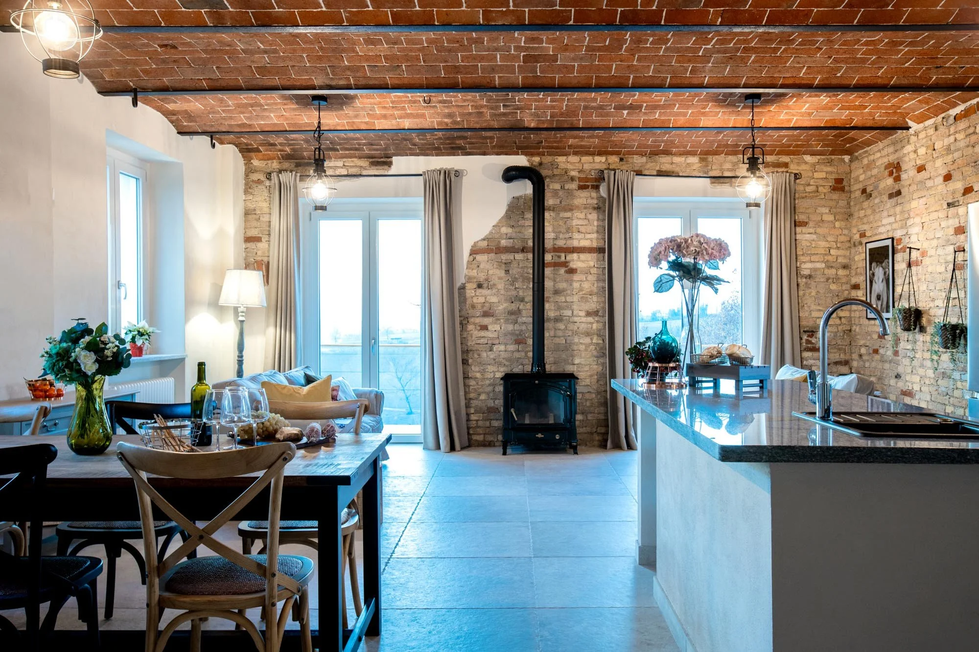Interior of a elegant and modern kitchen and dining area featuring exposed brick walls and ceiling beams, large windows with curtains, a kitchen island with a sink, a wood-burning stove, and a dining table decorated with flowers and wine glasses.
