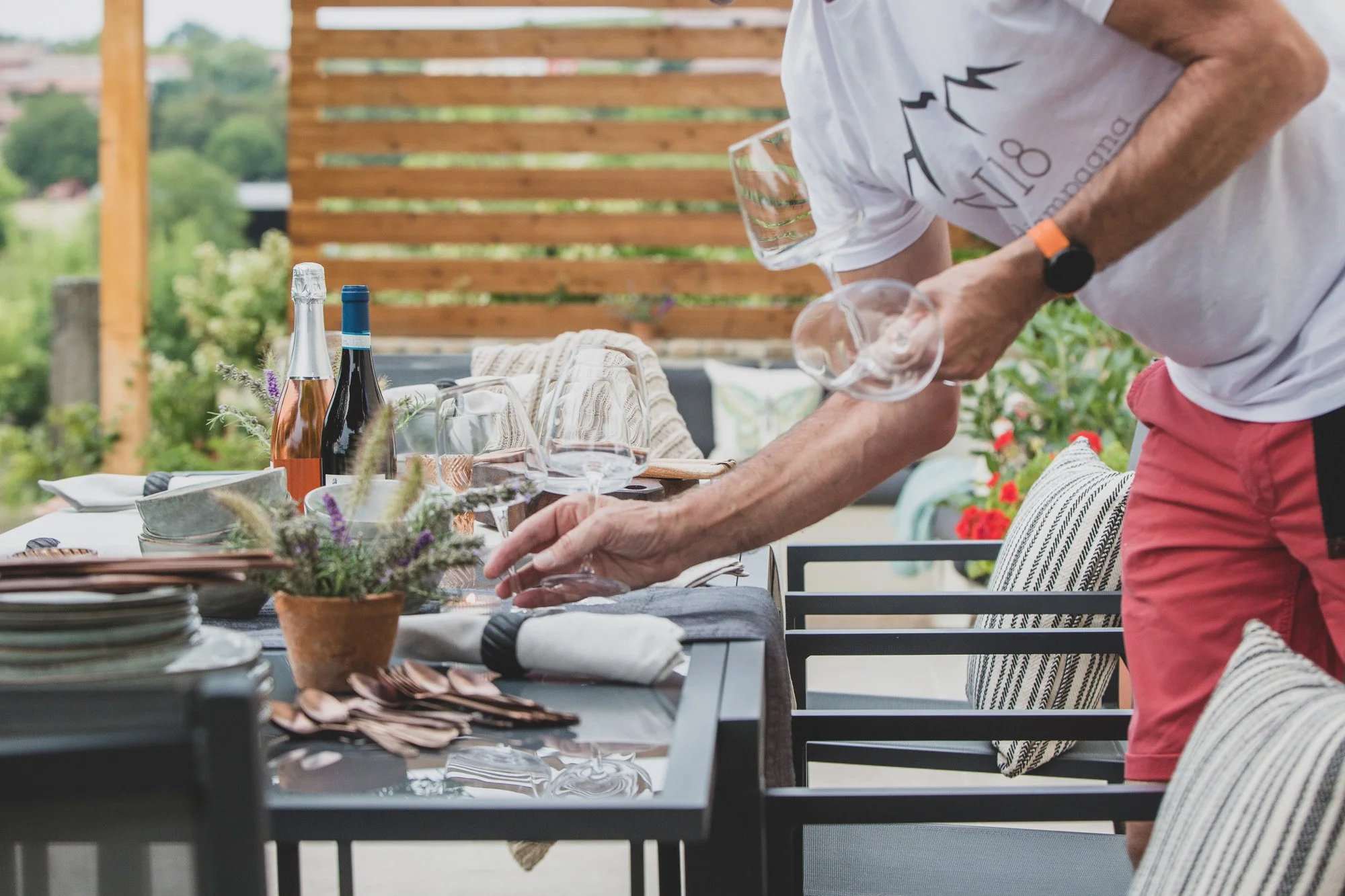A person setting a dining table outdoors with a sunset view, decorated with wine bottles, glassware, and a potted plant, on a patio with a wooden privacy screen in the background.