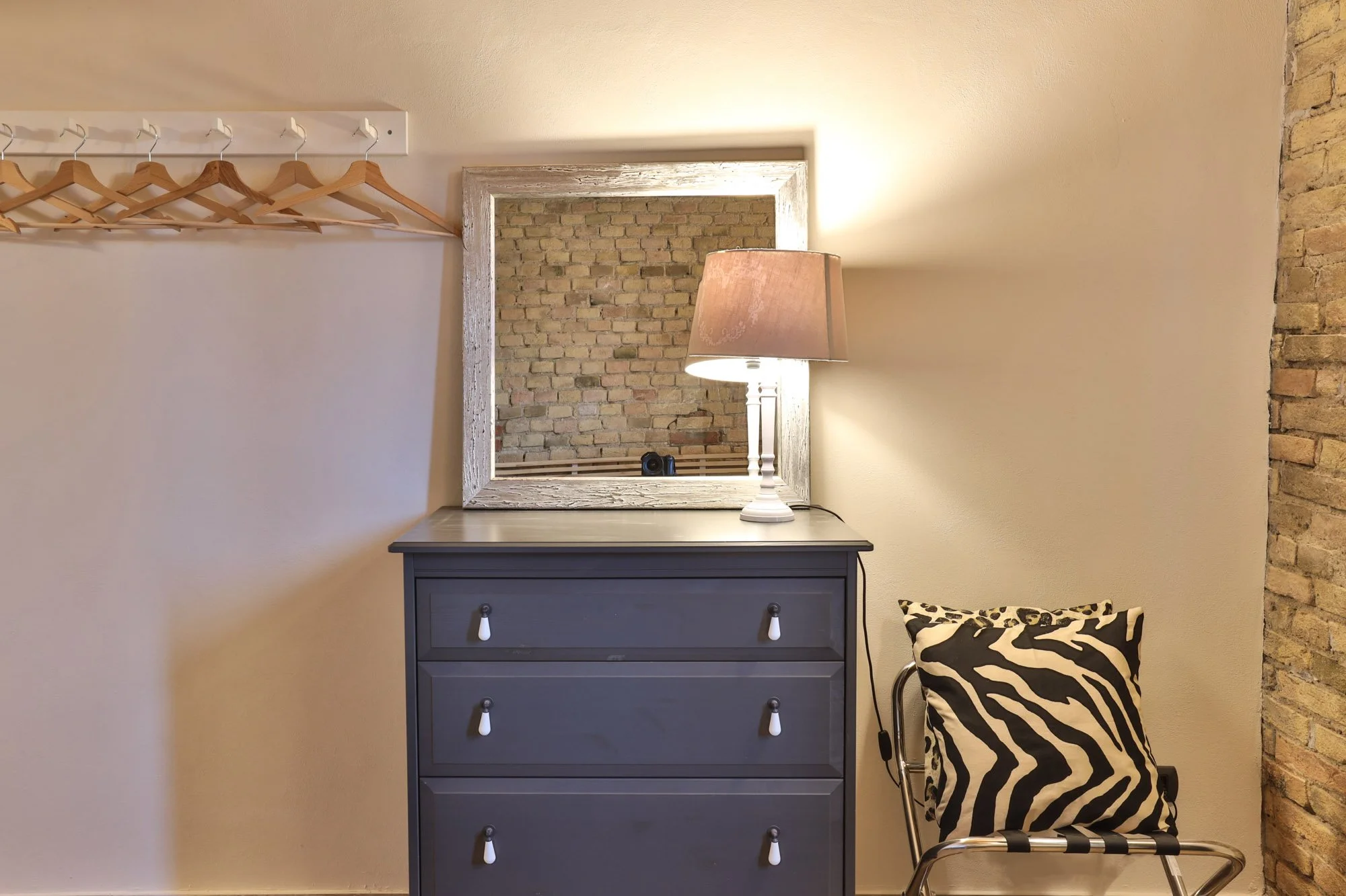 Interior of a room with beige walls, exposed brick accent wall, and a white framed mirror. There is a blue dresser with three drawers and white handles, a table lamp with a pink shade, a black and white zebra pattern cushion, and a metal-framed chair