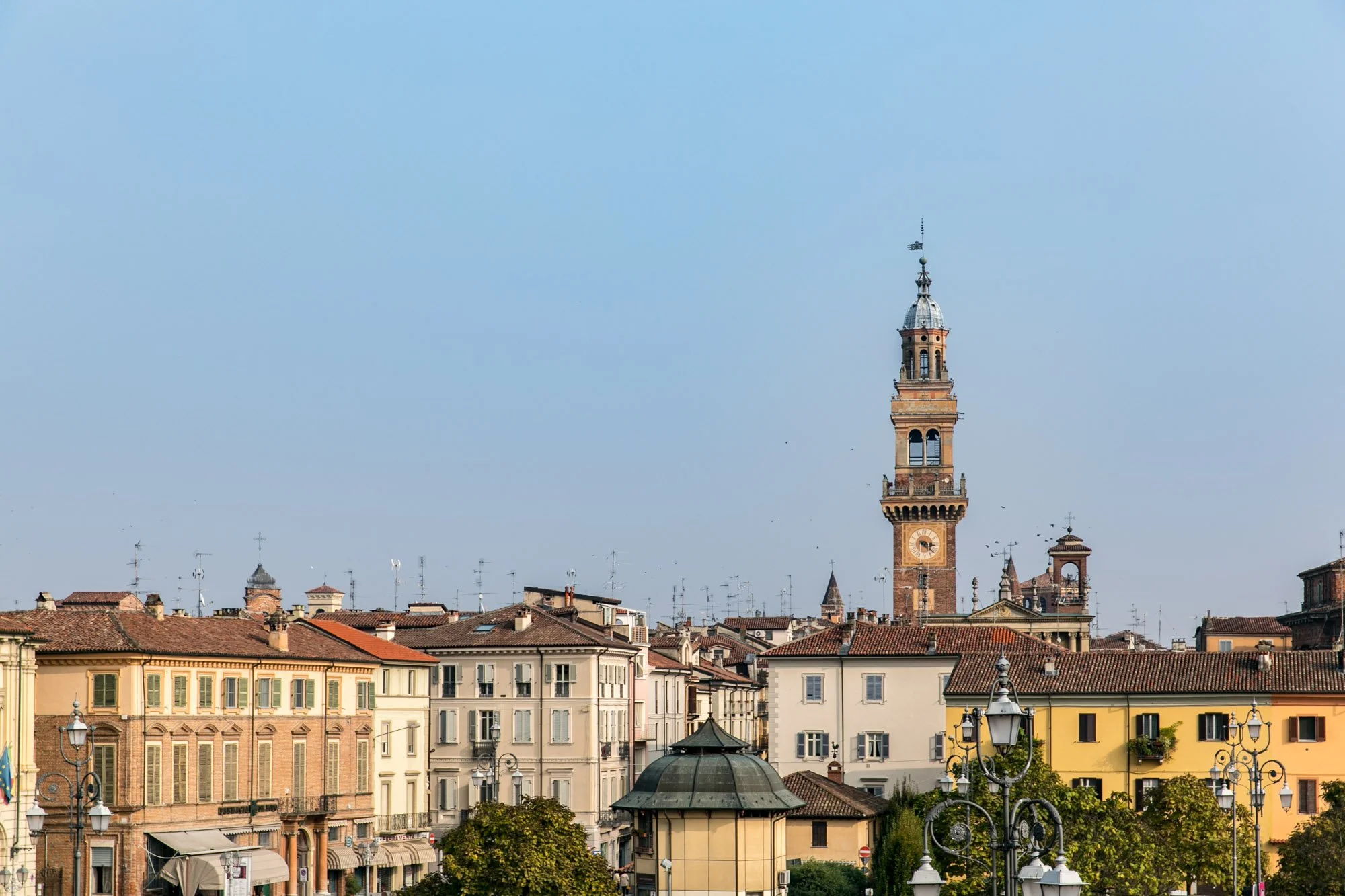Cityscape view of historic buildings  in Asti with a tall clock tower in the centre topped with a weather vane against a clear blue sky.