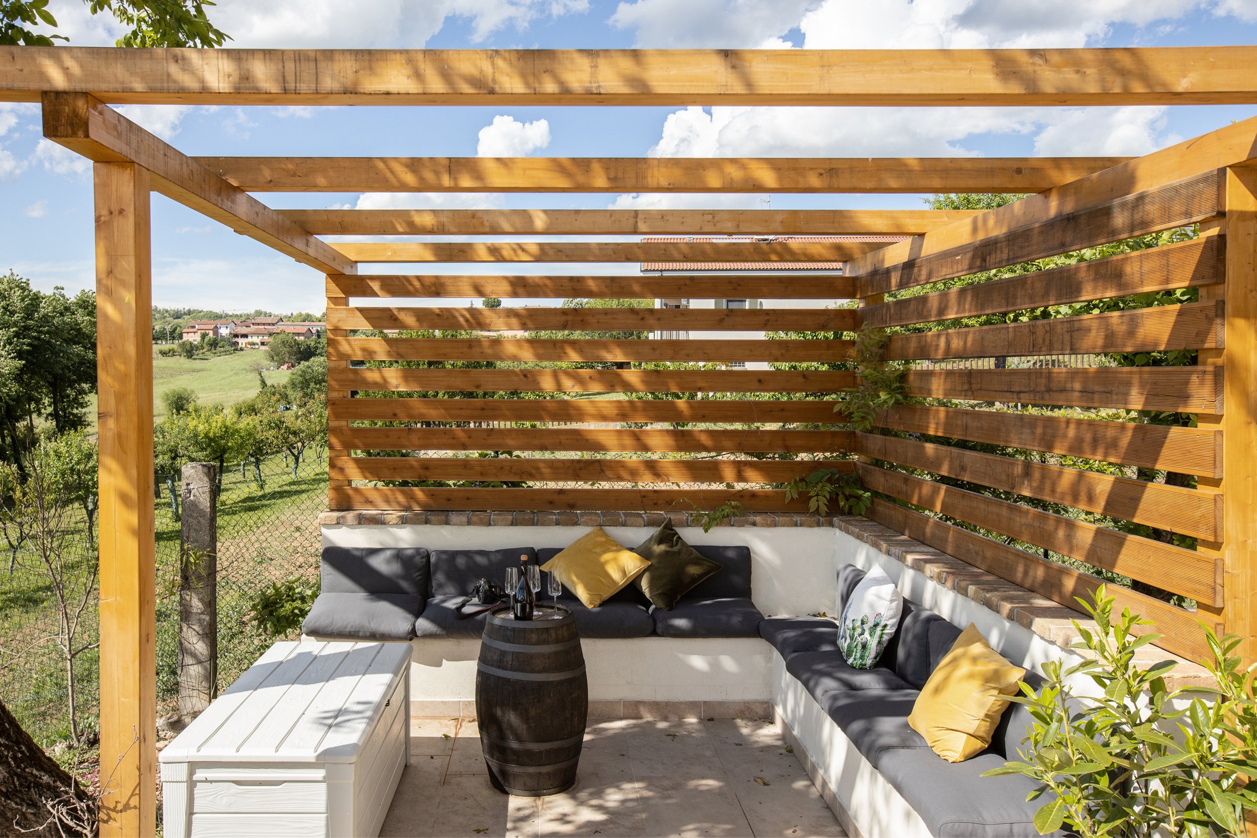 Outdoor seating area with a black sectional sofa decorated with gold pillows, a barrel-shaped table with drinks, a white side table, and a wooden pergola with horizontal slats, overlooking a green landscape with trees and houses under a partly cloudy