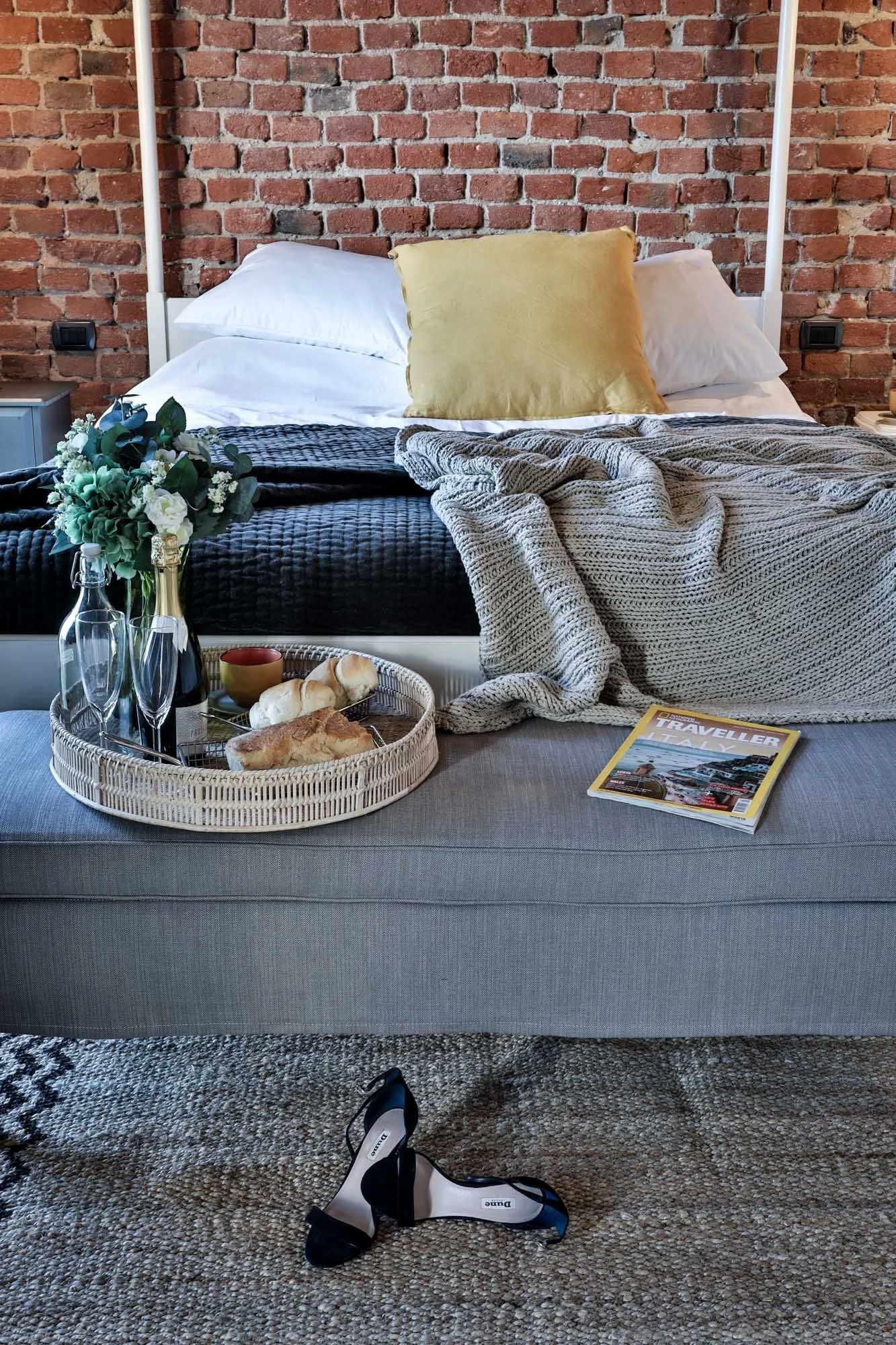 A bedroom with an exposed brick wall, a bed with white pillows, a yellow pillow, and a gray blanket. On the bed, there are a bouquet of flowers, a magazine titled 'Traveller Italy,' and a tray with bread, cheese, and a small bowl. A pair of black and