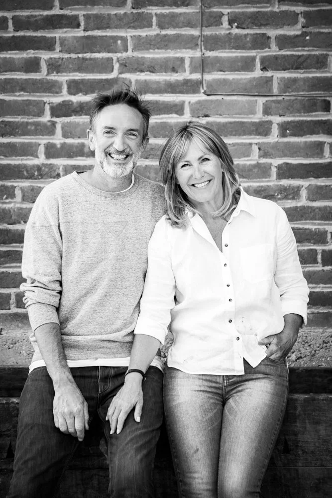 A black-and-white photo of a smiling man and woman sitting close together against a brick wall, casually dressed, with the woman resting her hand on her lap.