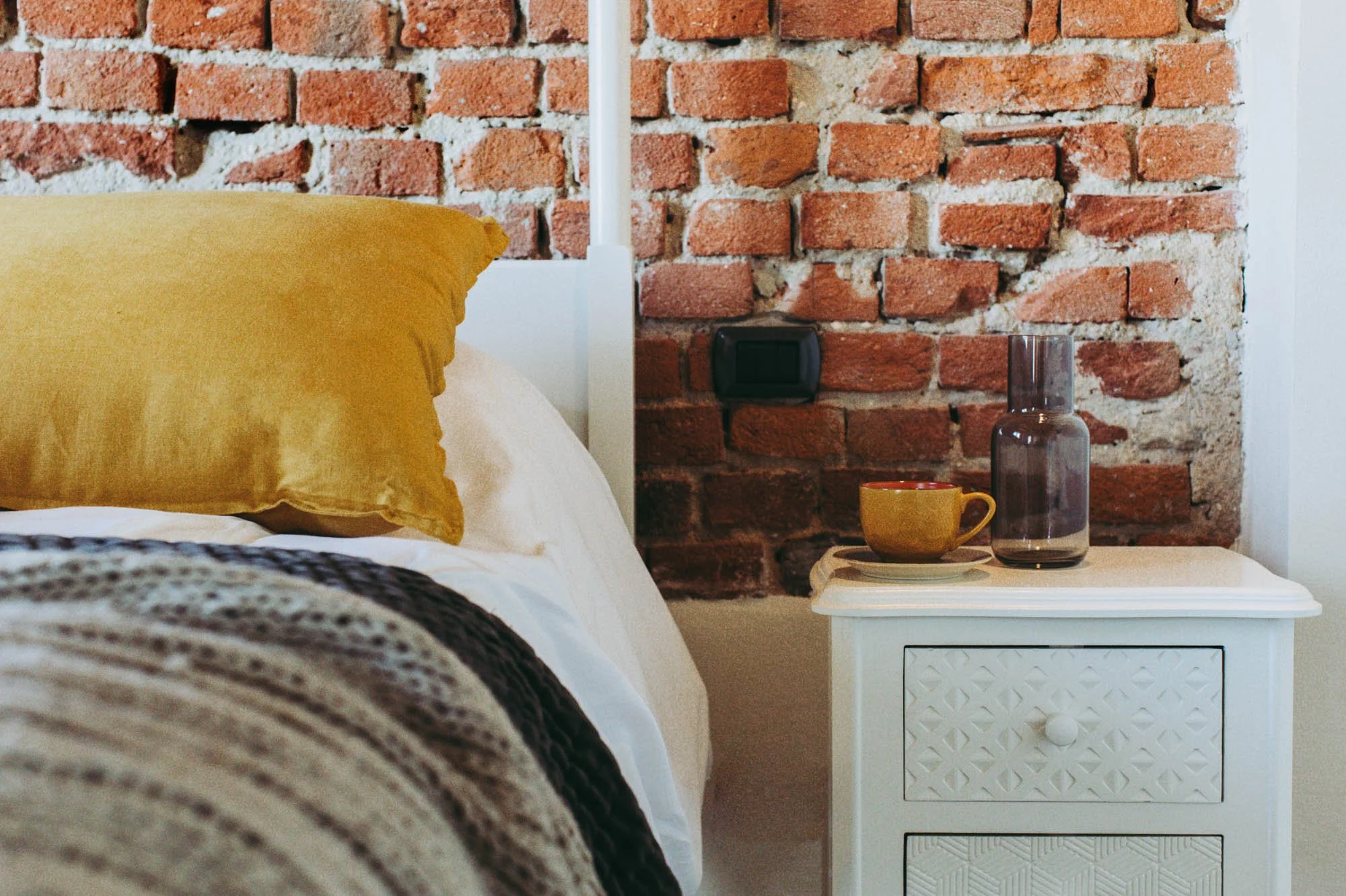 A bedroom with a large yellow pillow on the bed, a white nightstand with a yellow mug, a glass water bottle, and a tray. The bed has a textured beige and gray blanket. The background features an exposed red brick wall and a white nightstand with a dr