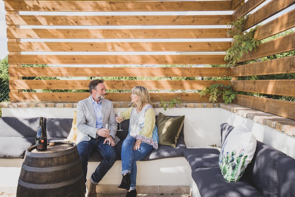 A man and woman sitting on a cushioned outdoor patio lounge, sharing a glass of wine, with a wooden privacy fence and greenery in the background.