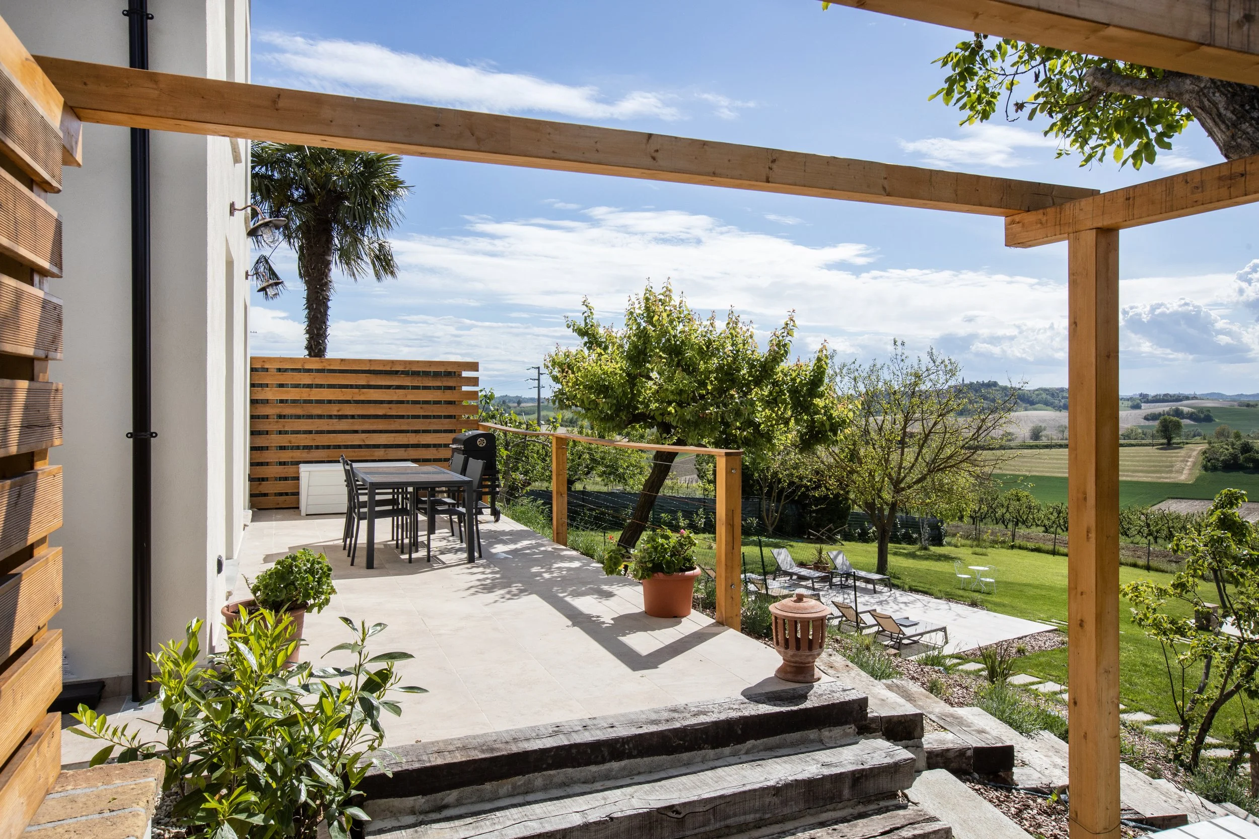 Residential patio with outdoor dining set, potted plants, and a scenic view of green fields, trees, and a bright blue sky with clouds.