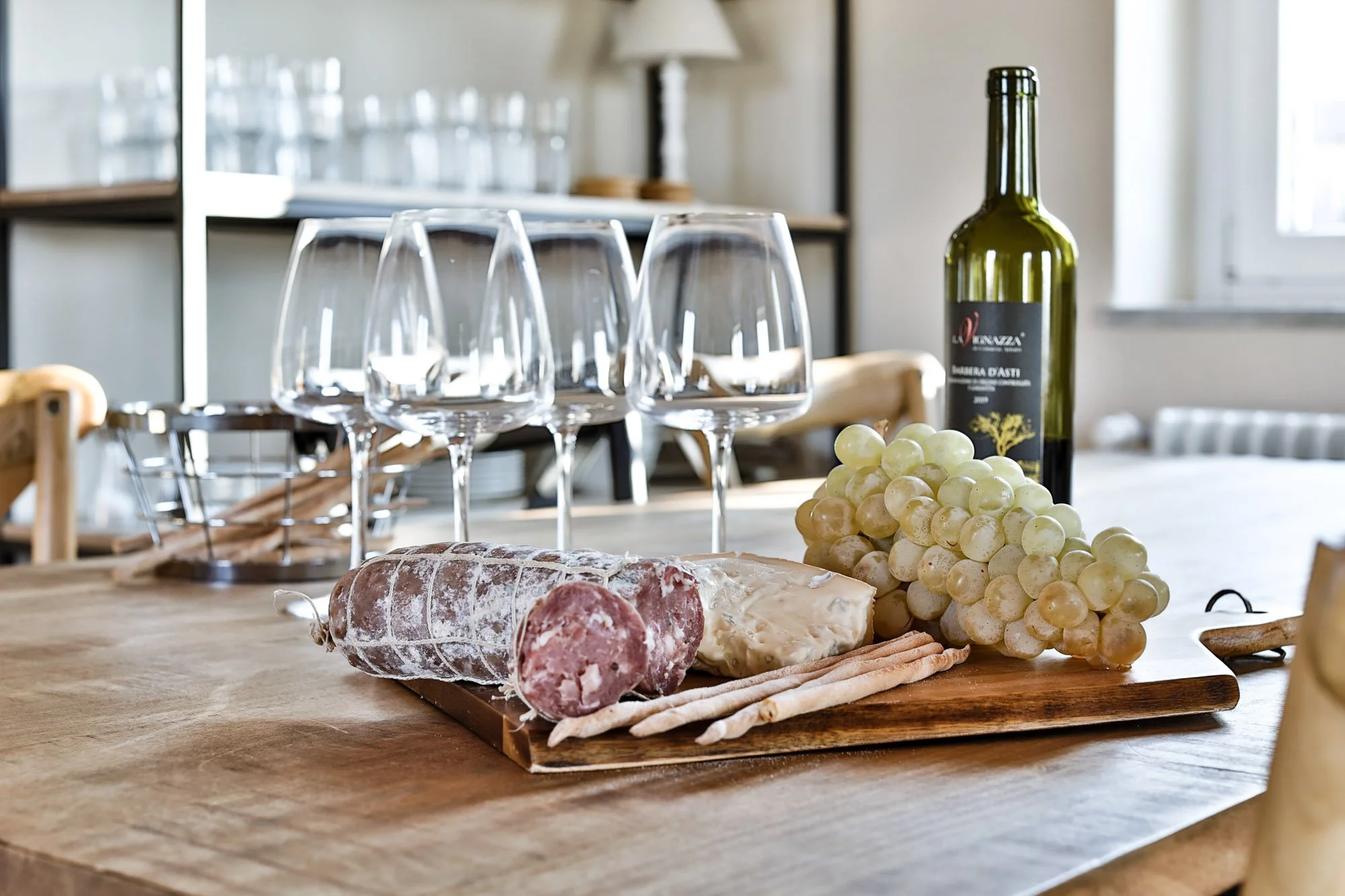 A wooden table with a bunch of grapes, slices of cured meats, and a loaf of bread, with wine glasses, a wine bottle, and a lamp in the background.
