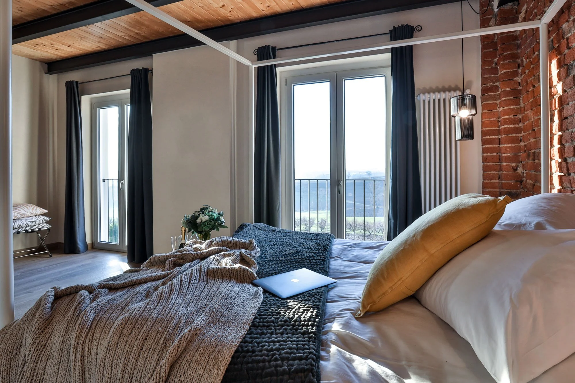 A bedroom with an exposed brick wall, large windows with black curtains, and a wooden ceiling. The bed has yellow pillows, a laptop, and a cozy blanket. There is a flower arrangement on a table near the window.