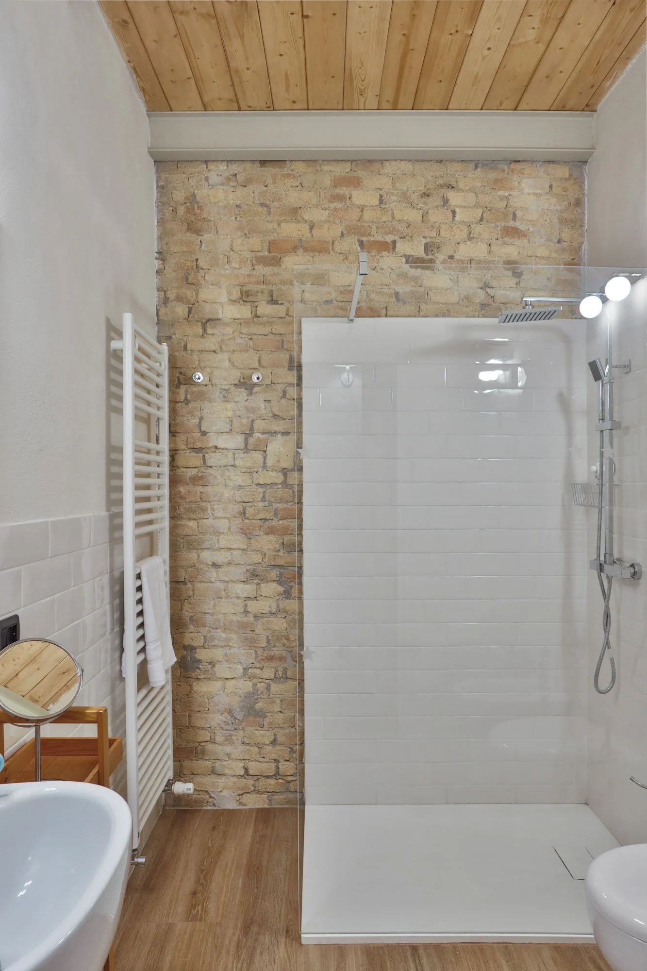 Modern bathroom with exposed brick wall, wood ceiling, white tiles, shower area with rainfall showerhead, heated towel rail, and wooden furniture.