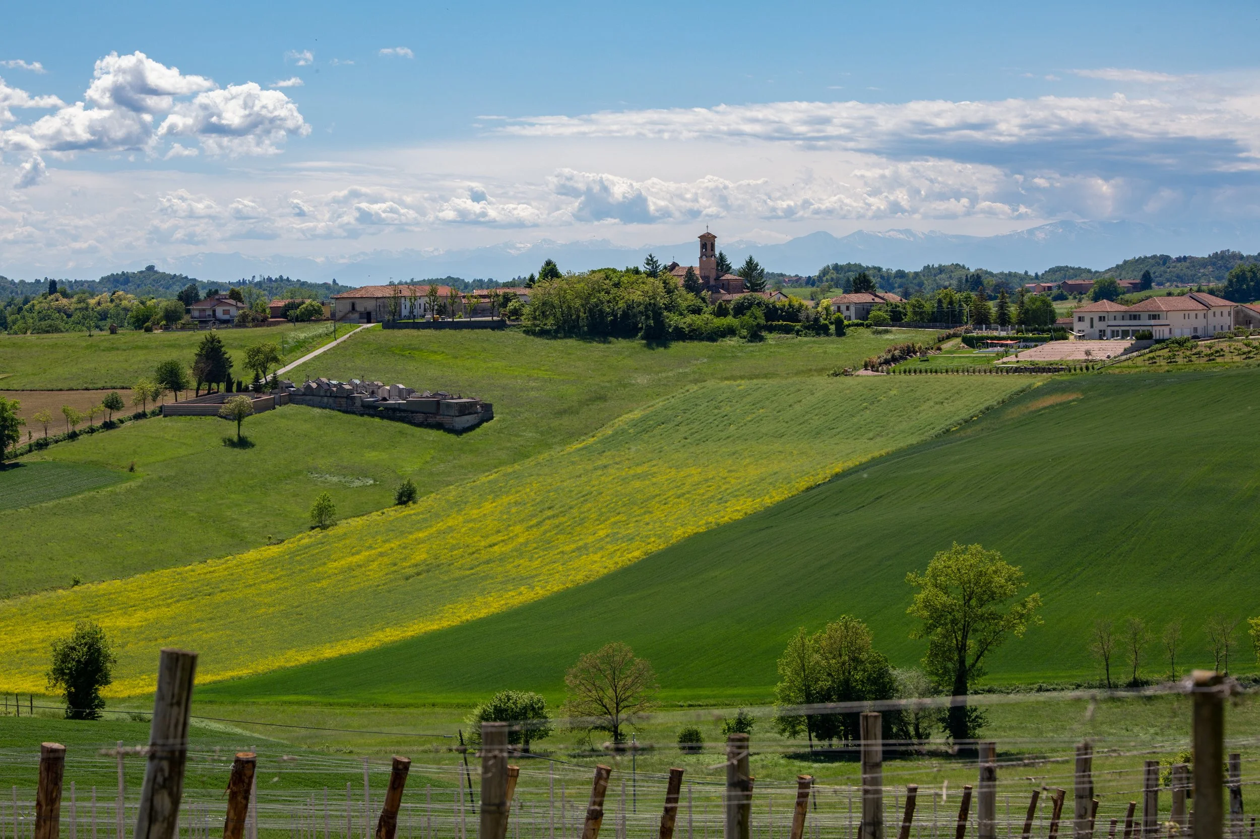Scenic landscape of rolling green hills with scattered trees, farmhouses, and a church, under a partly cloudy sky with distant mountains.