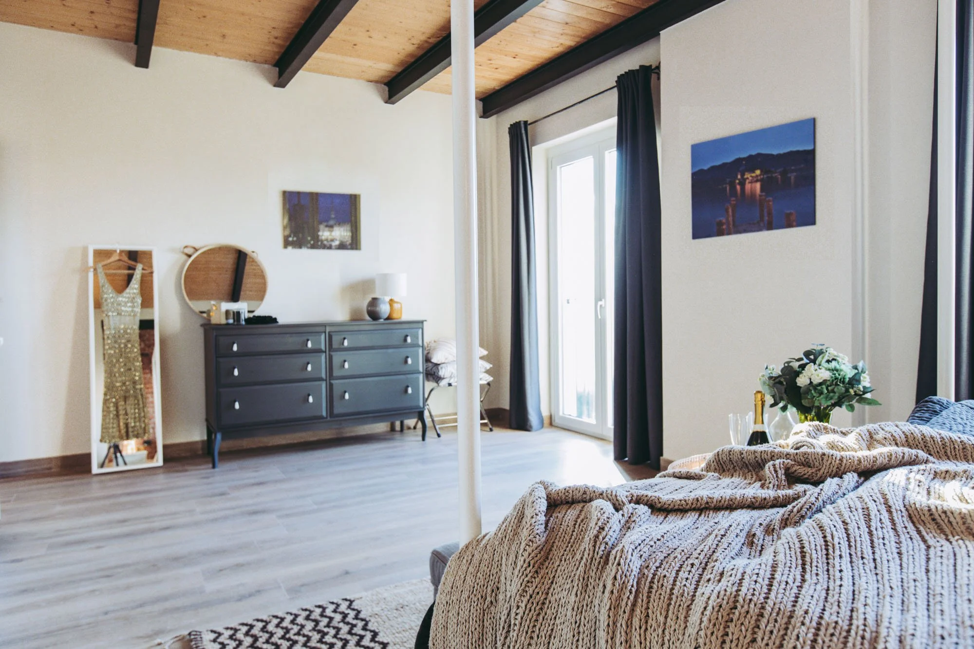 A cozy bedroom with a wooden ceiling, white walls, and large glass doors opening to a balcony. A gray dresser with a lamp and decorative items is against the wall, and a full-length mirror is leaning beside it. The bed has a chunky beige knit blanket