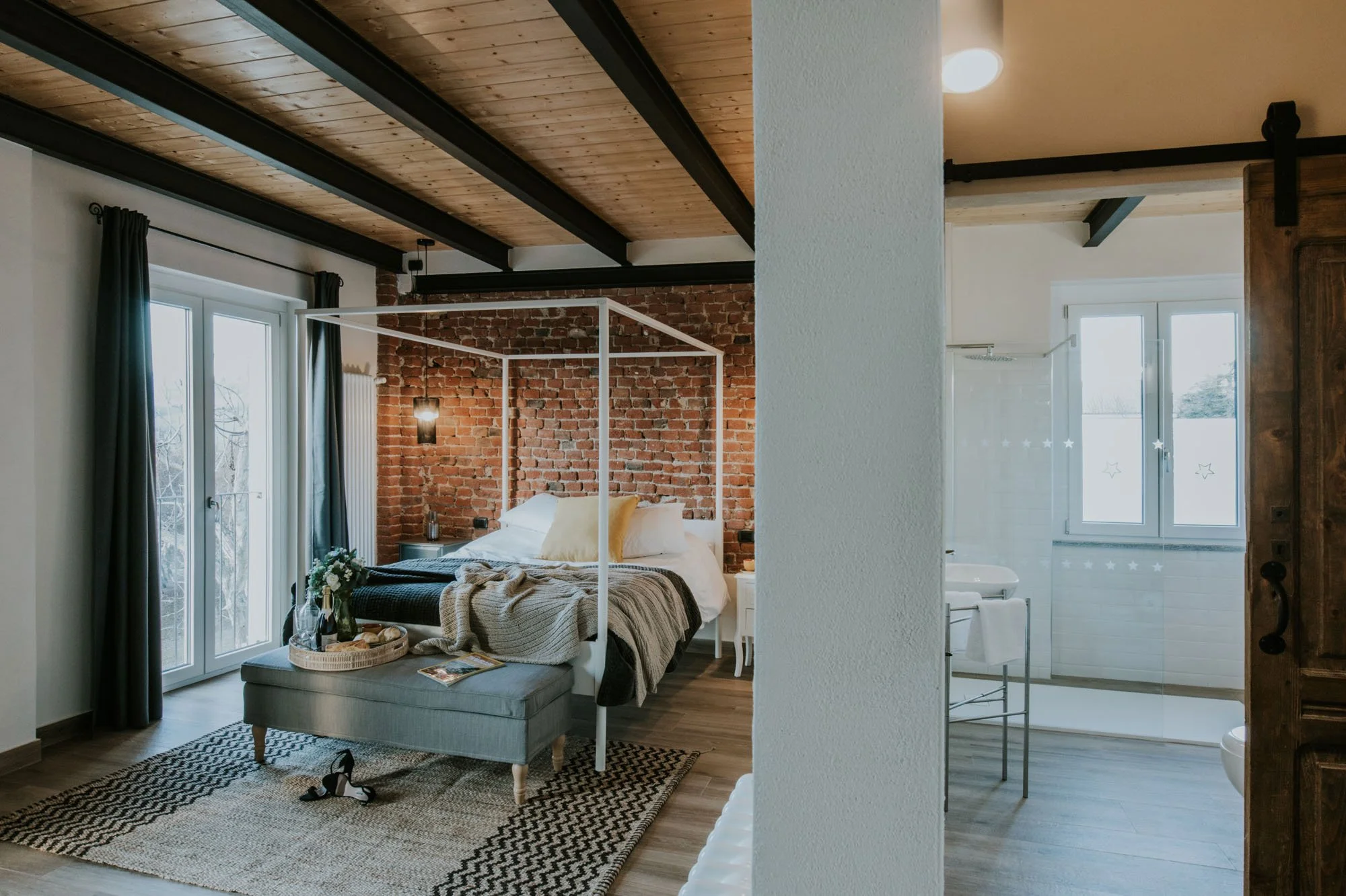 A cozy bedroom with a four-poster bed, a beige blanket, a gray upholstered bench, and a tray with snacks. Large windows with dark curtains, brick accent wall, wooden ceiling with black beams, and a partial view of a bathroom with a window and white t