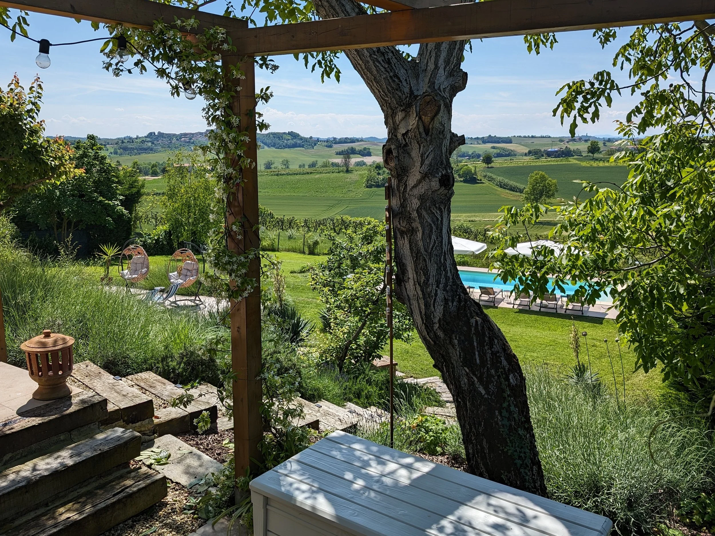 A backyard view with a large tree, garden furniture, and a swimming pool, overlooking rolling green hills and a blue sky.
