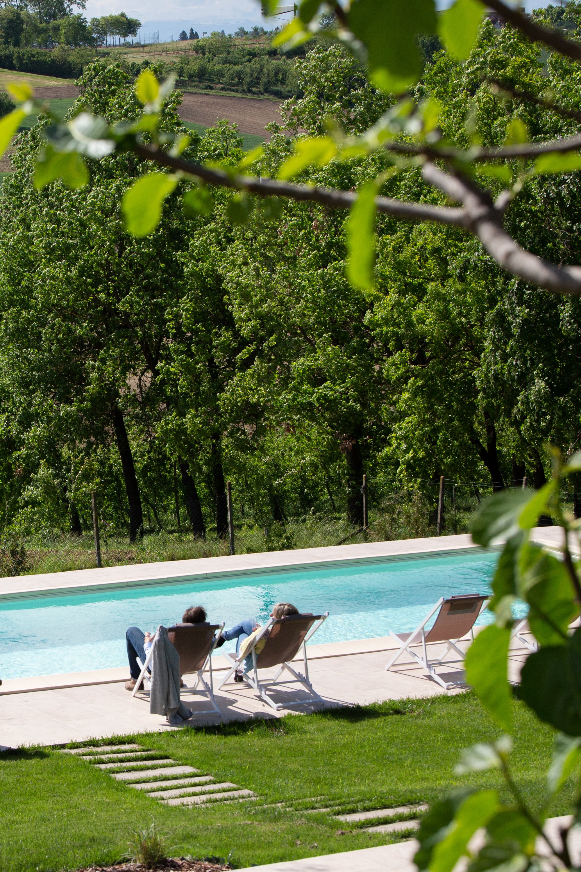 People relaxing on lounge chairs by a swimming pool with green trees and hills in the background.