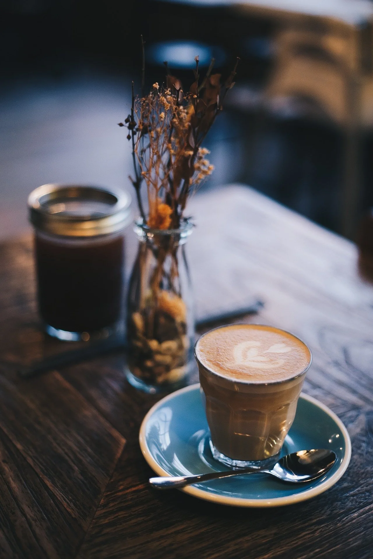 A Bicerin art coffee in a glass cup on a saucer with a spoon, a small jar, and a dried flower arrangement on a wooden table in a cozy Turin café.
