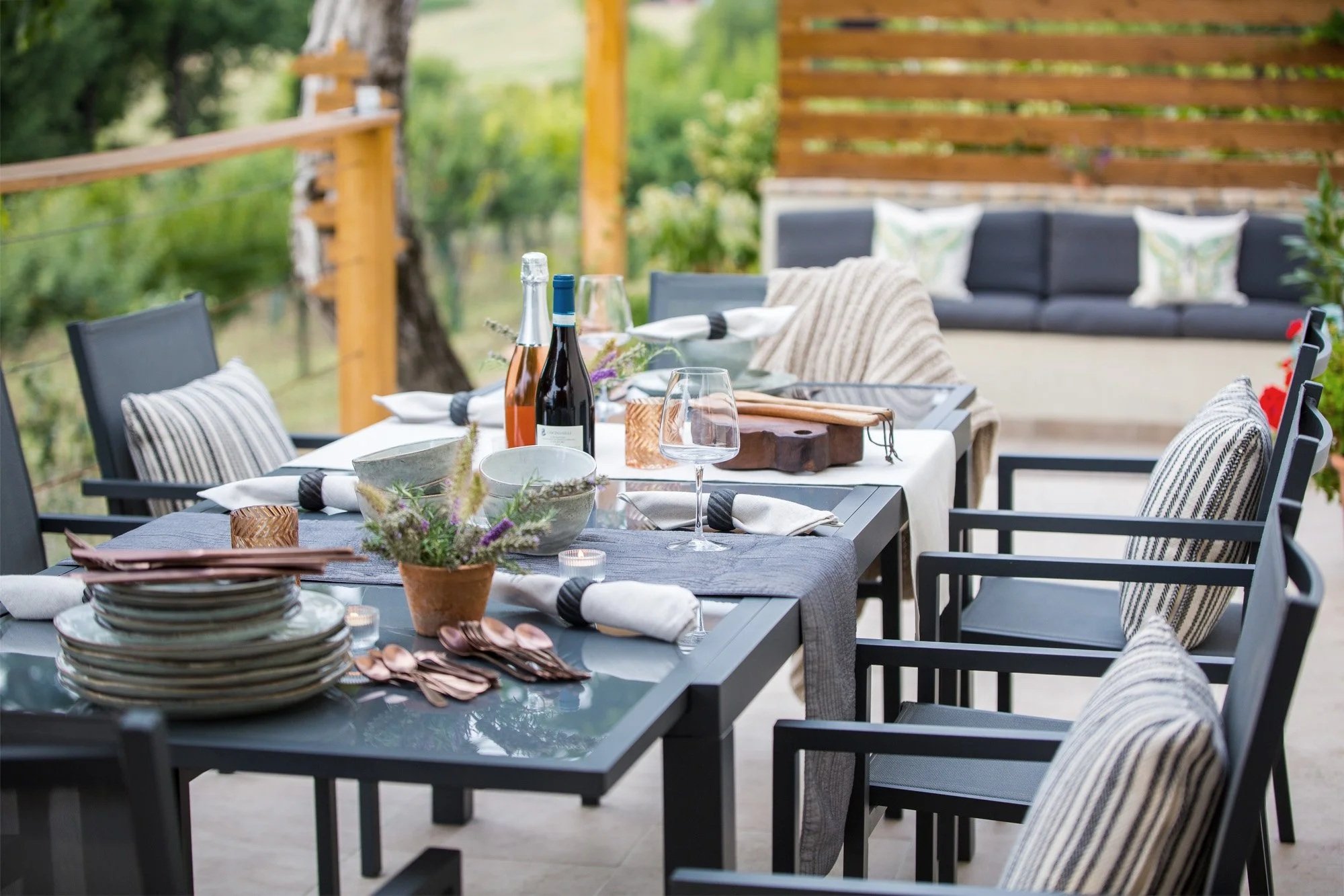 Outdoor dining table set with plates, glasses, cutlery, wine bottles, and flowers in a cozy backyard with a wooden fence and greenery.