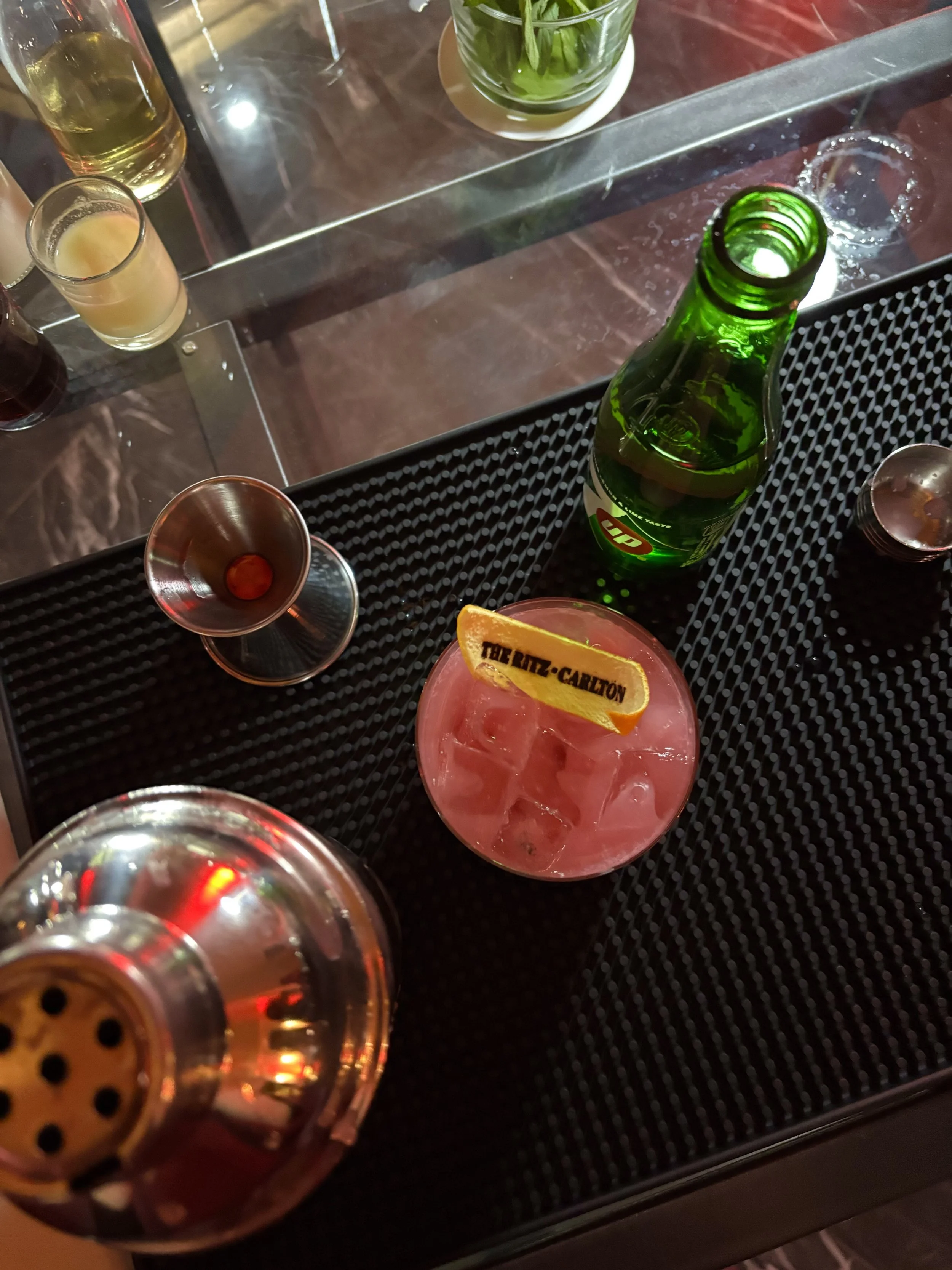View of a bar table with various drinks, including a green bottle of beer, a glass of orange-colored drink with ice and a lemon wedge, and a cocktail garnished with an orange slice labeled 'TEL RIZ-CARLTON.'