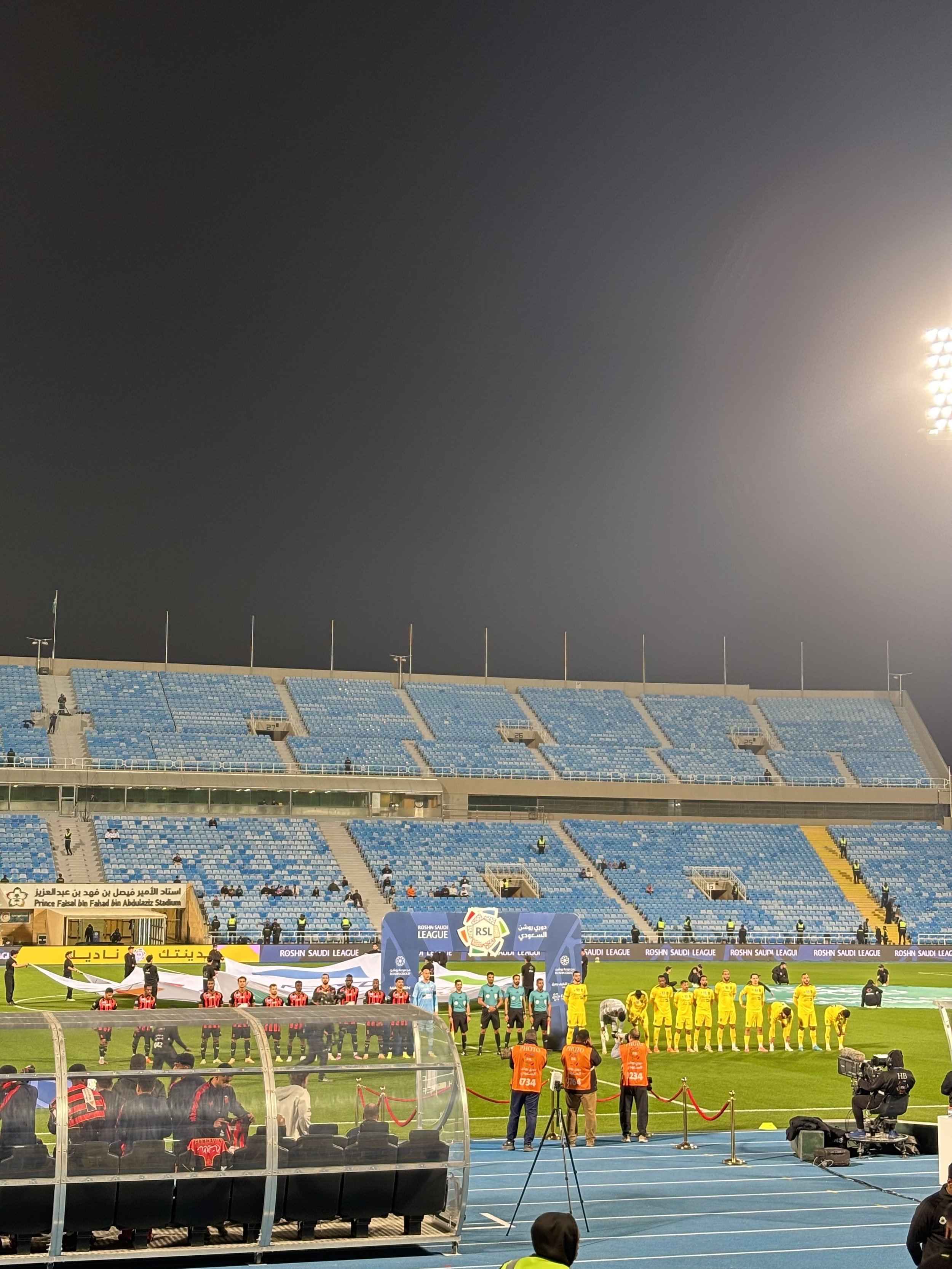 A stadium with blue and yellow seating, illuminated by bright stadium lights, and players lined up on the field for a soccer match. There are officials and cameras set up, with banners that read 'ROSHN SAUDI LEAGUE' and other Arabic text.
