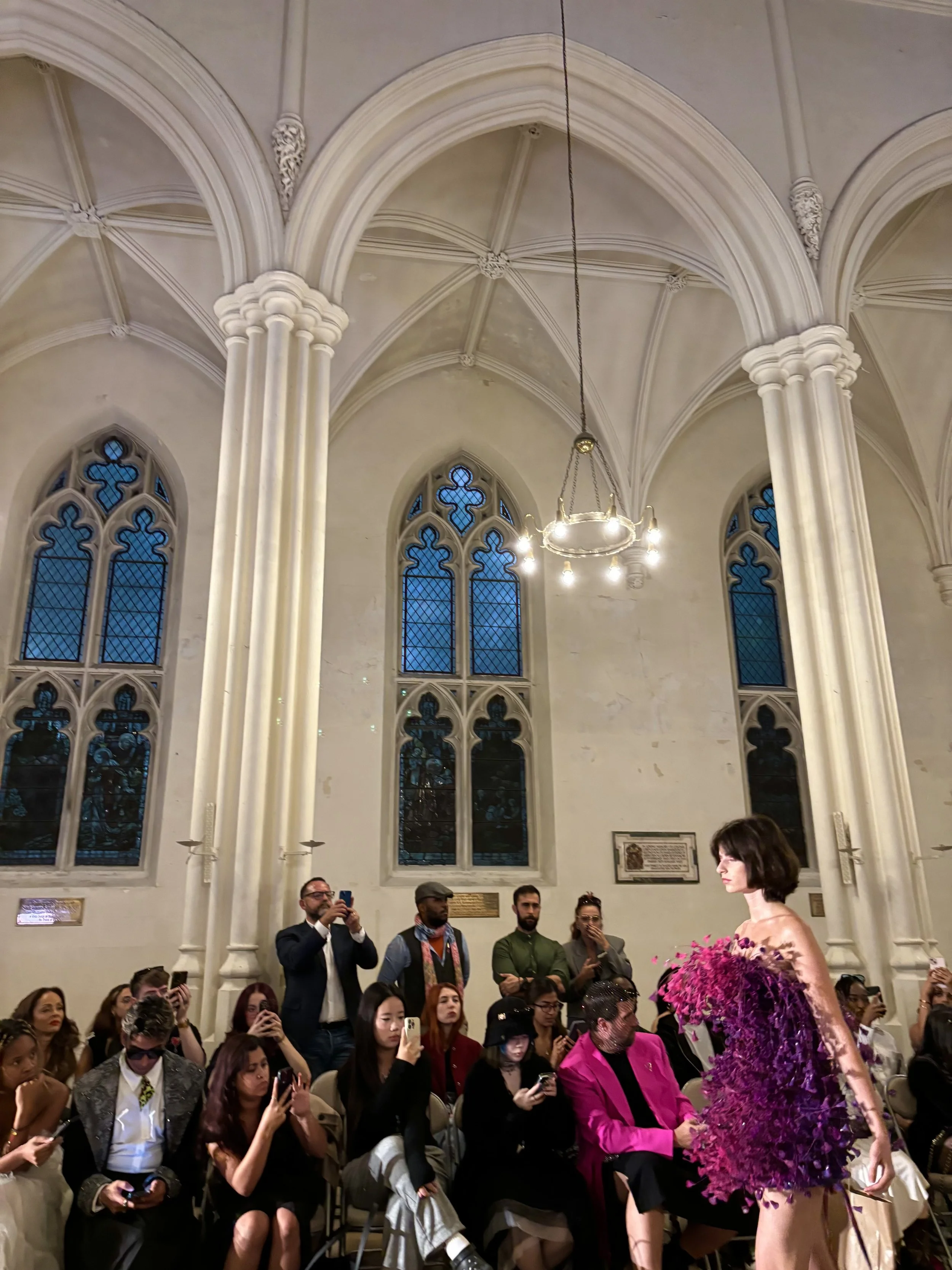 People attending London Fashion show inside a church with tall arched windows and ornate ceiling details. A model with styled hair and elaborate purple dress is in the foreground, while audience members are seated and standing, some taking photos.