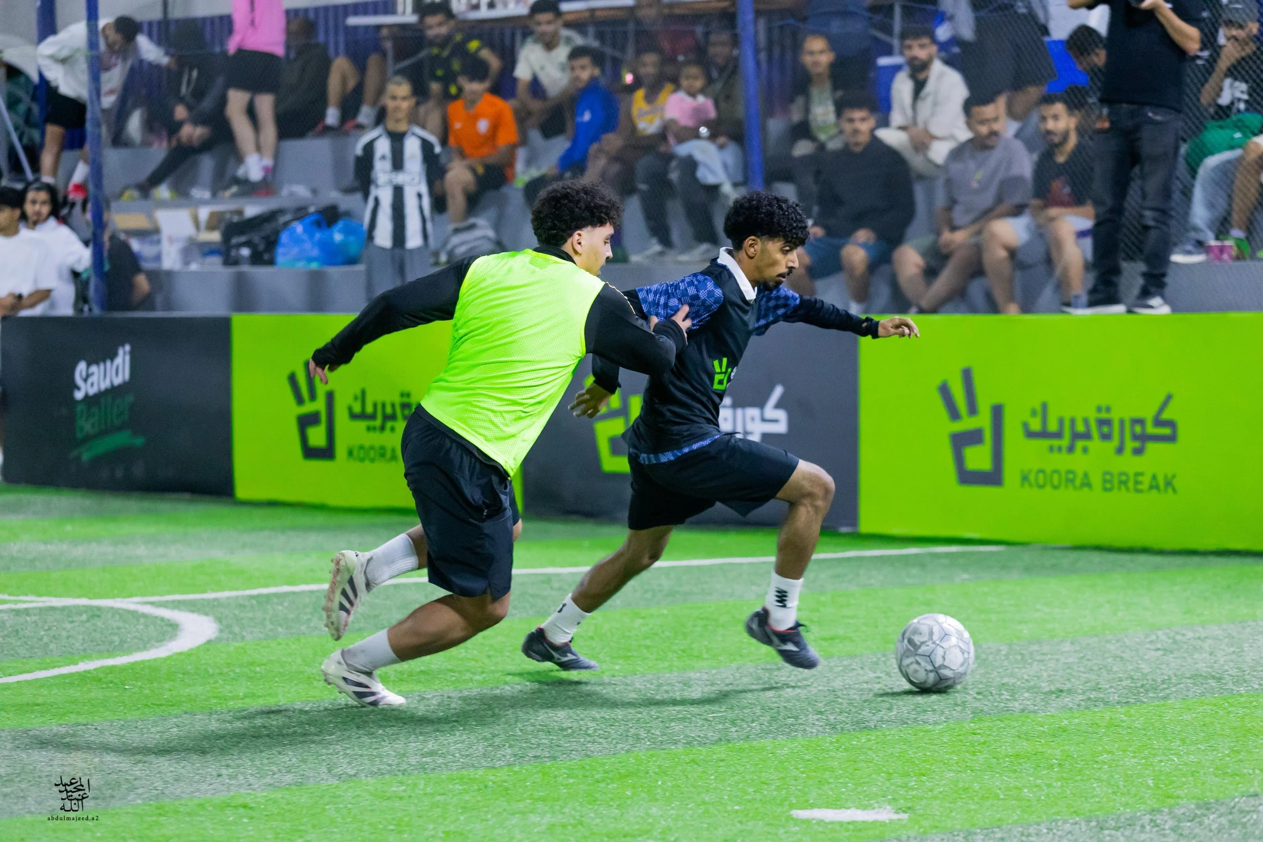Two soccer players with curly hair running on a green field towards a soccer ball. One player wears a neon yellow vest, the other wears black and blue sportswear. Spectators watch from the stands behind a blue fence.
