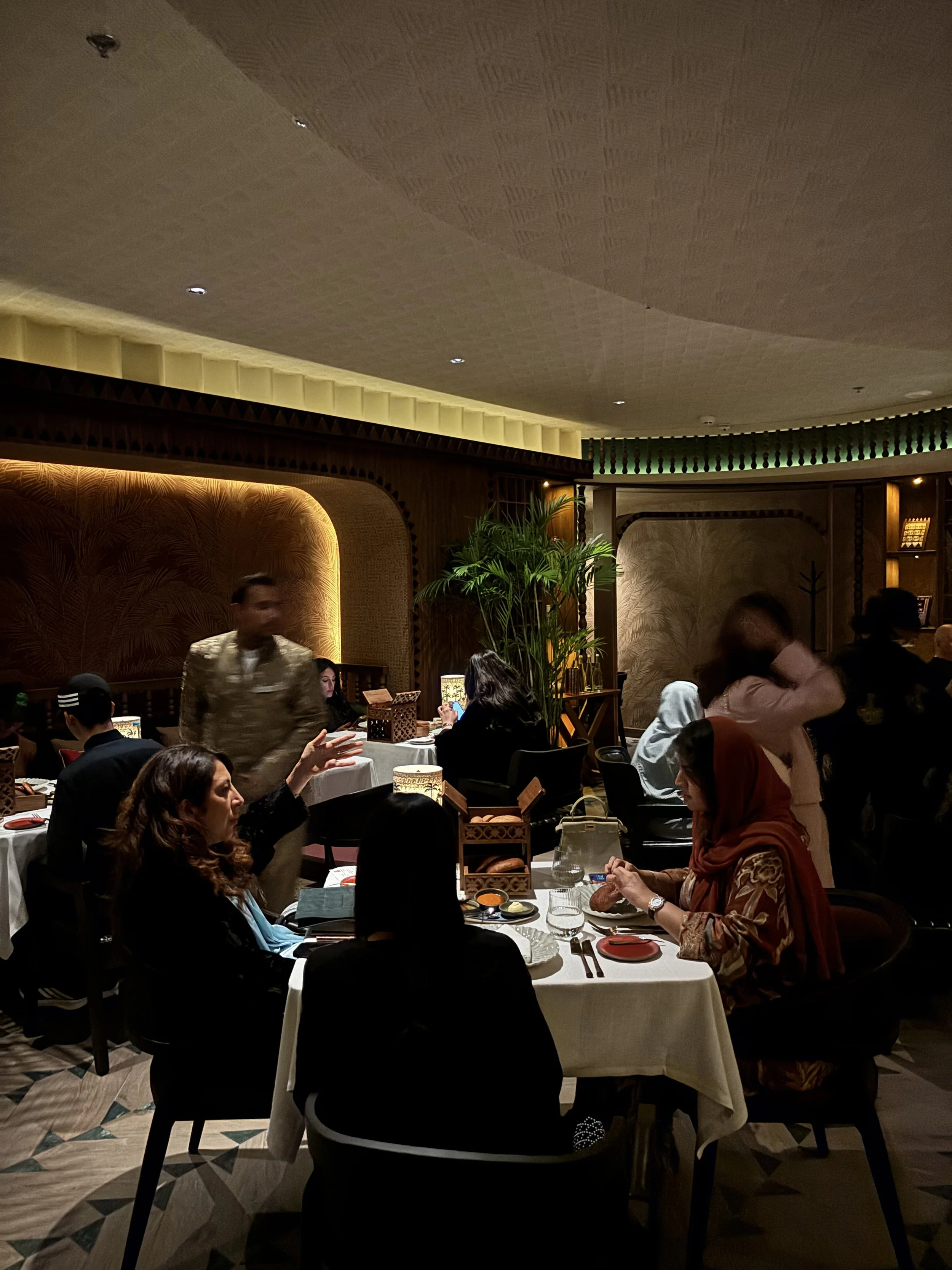 A crowded restaurant with people dining at tables, some of whom are talking, a waiter walking by, a woman eating, and the interior featuring warm lighting, decorative walls, and potted plants.