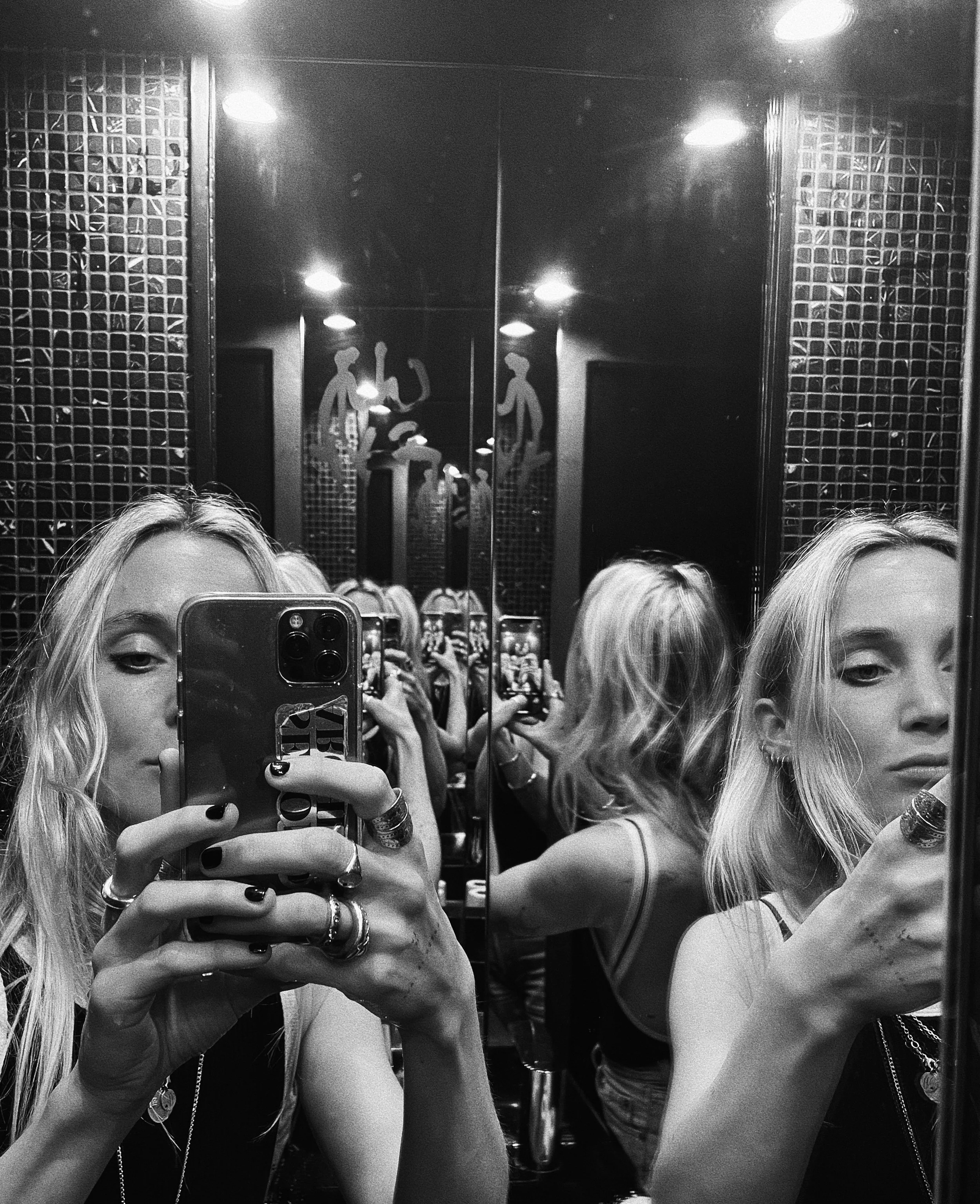 Black and white photo of two young women taking a selfie in a mirror. Both are holding phones, with the woman on the left wearing rings and bracelets, and the woman on the right wearing a necklace. The background features a decorative wall with female silhouettes and reflective surfaces.