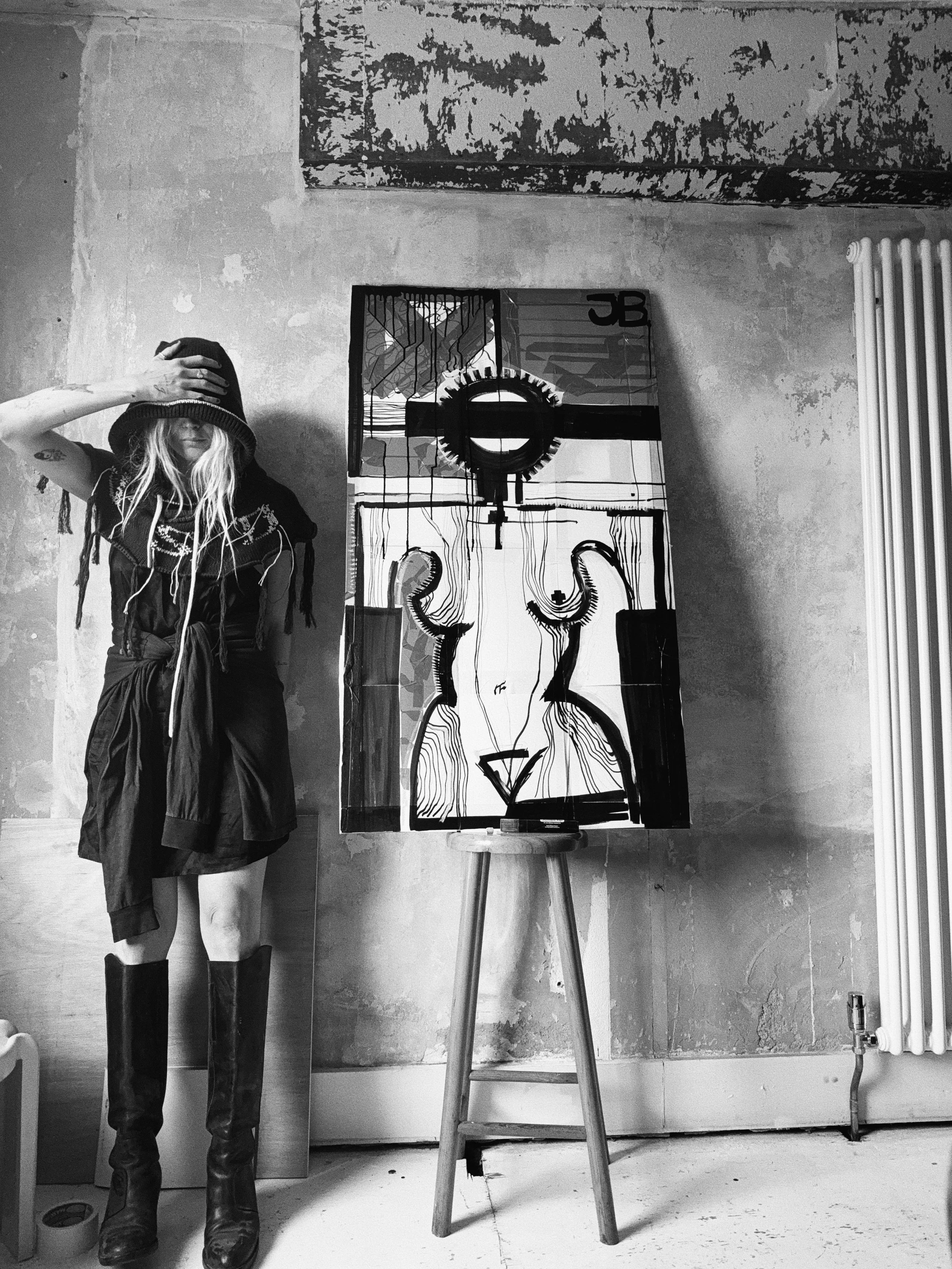 A woman wearing a hoodie with her face partially hidden, standing next to an easel with a modern, abstract painting. The setting appears to be an artist's studio with a textured wall, a radiator on the right, and a visible pipe on the floor.
