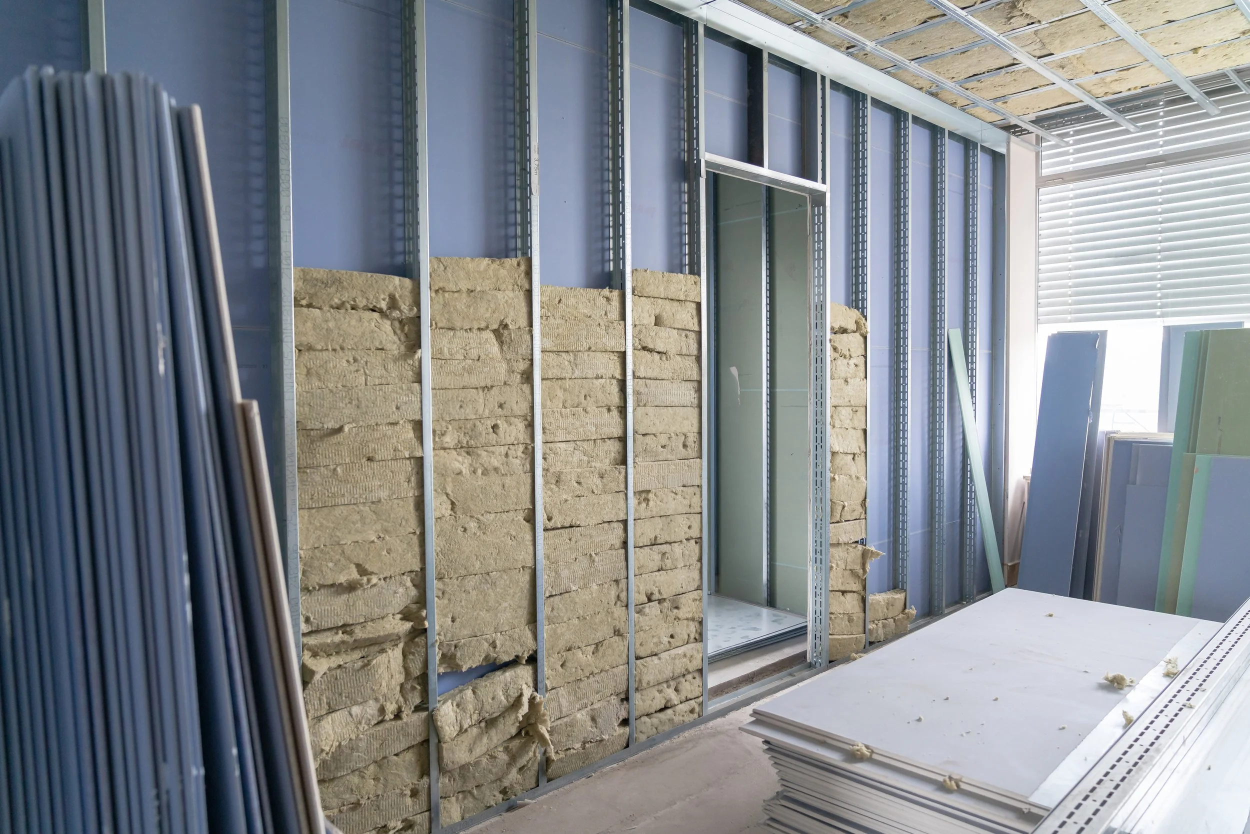 Innenansicht eines Raumes unter Bauarbeiten, mit Metall-Wandständern, Dämmmaterial und Baumaterialien auf dem Boden.