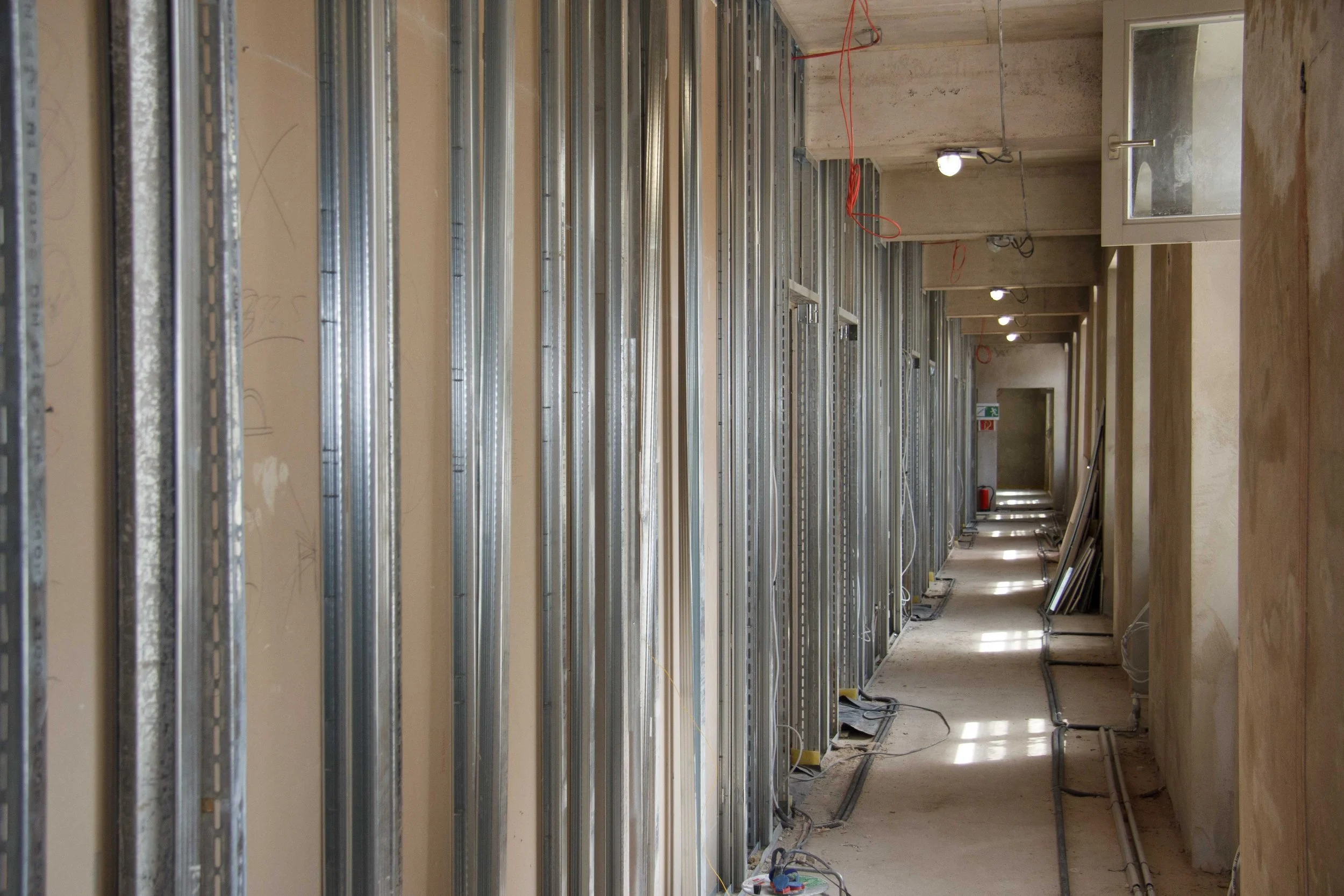 Innenansicht eines Gebäudes im Bau, mit Metallrahmen für Wände und Deckeninstallation, Kabel hängen herab.