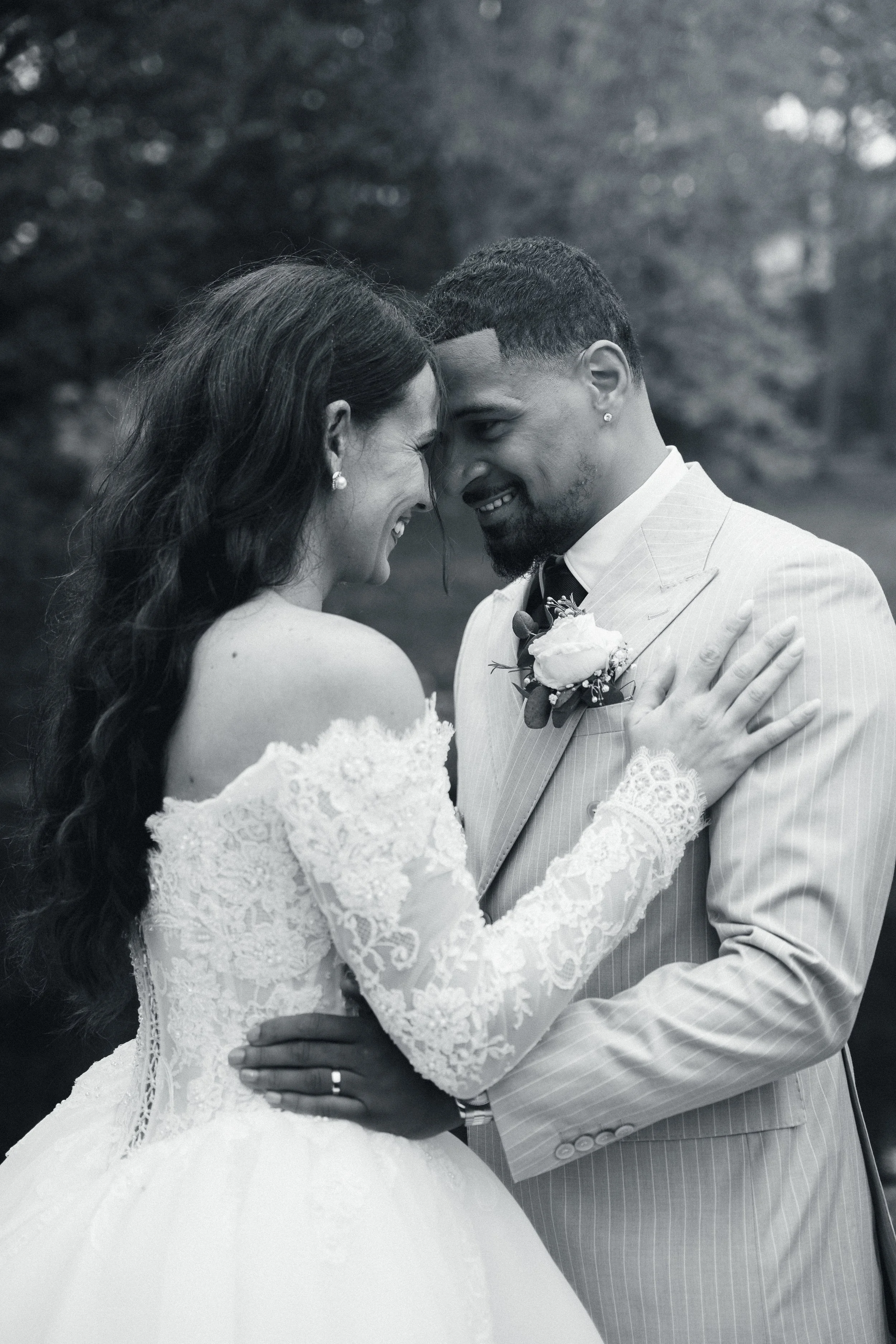 Black and white photo of a bride and groom touching their foreheads, smiling, and embracing each other outdoors during a wedding.