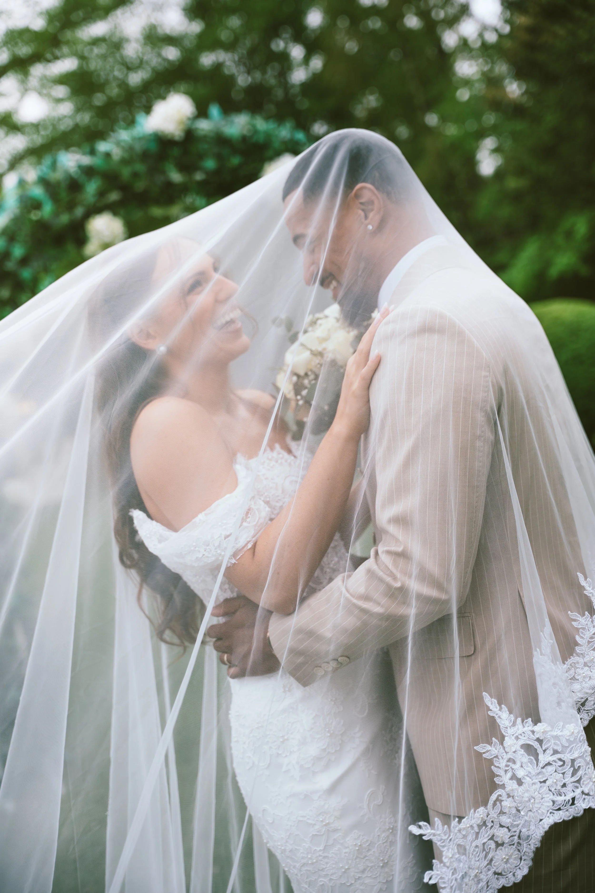 A newlywed couple standing close under a veil outside, smiling and looking at each other, with green trees in the background.