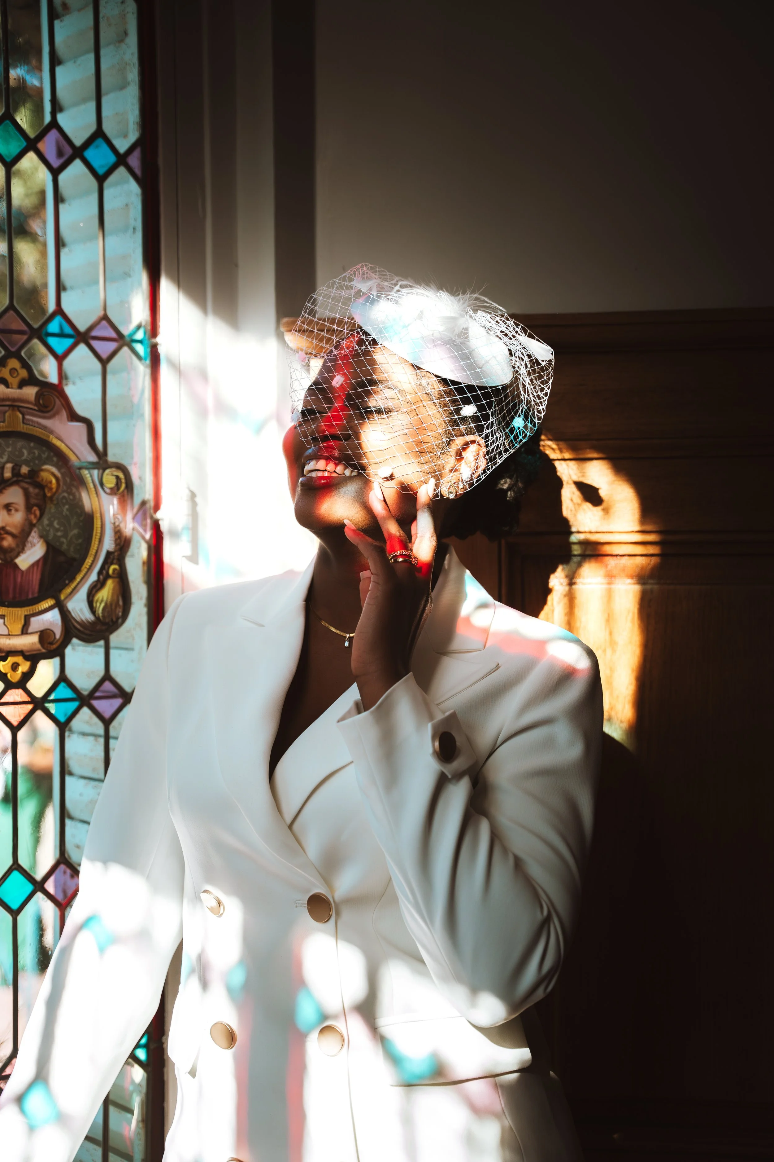 Femme souriante portant un manteau blanc avec un voile décoratif, à côté de vitraux colorés, éclairée par la lumière naturelle.