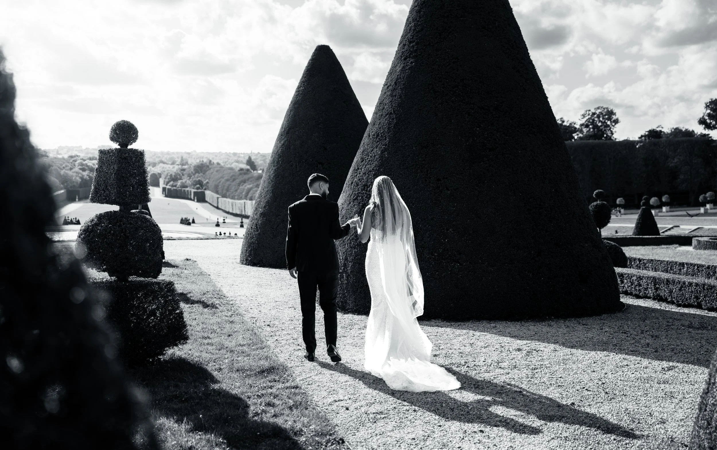 A black and white photo of a bride and groom holding hands and walking in a formal garden with large sculpted topiary trees.