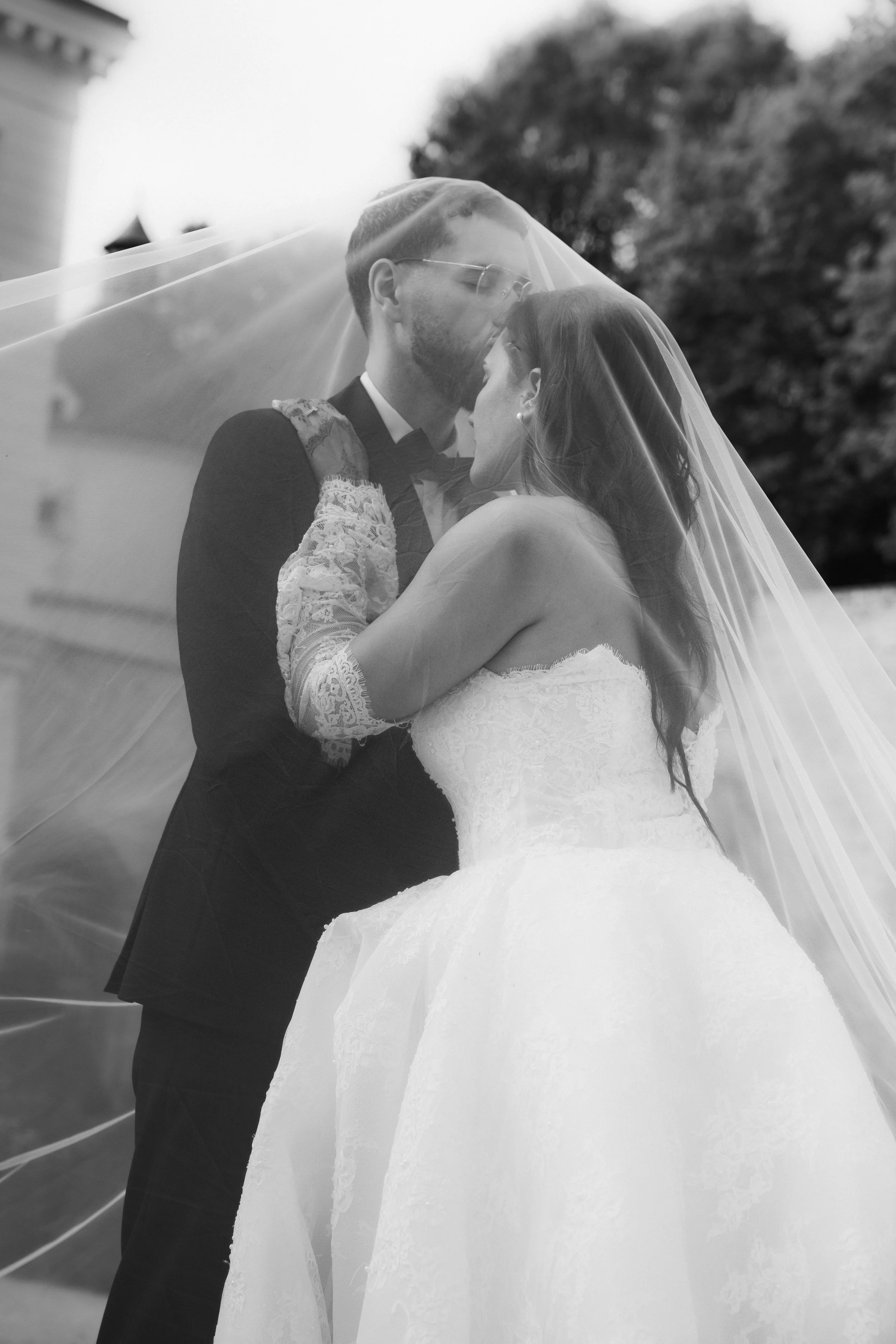 Une photo en noir et blanc d'un couple de mariés lors de leur mariage, partageant un moment intime sous un voile de mariée, avec des arbres en arrière-plan.