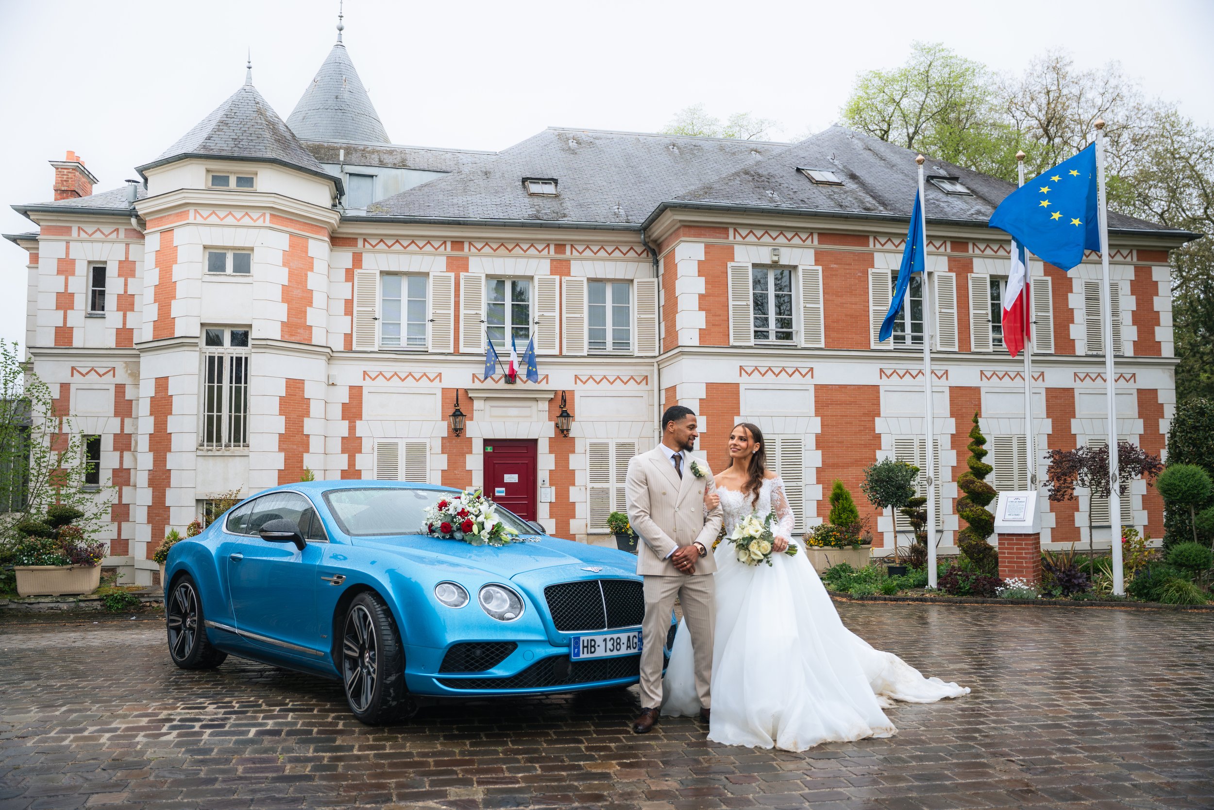 A newlywed couple in wedding attire standing in front of a blue sports car decorated with wedding flowers, parked outside a large brick and stone mansion with flags, trees, and cloudy sky in the background.