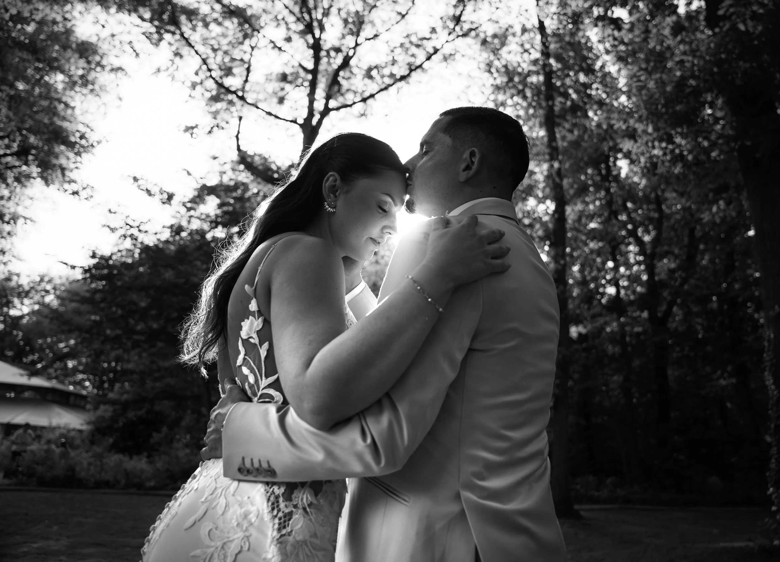 A black and white photo of a bride and groom embracing outdoors, with the groom kissing the bride's forehead, surrounded by trees.