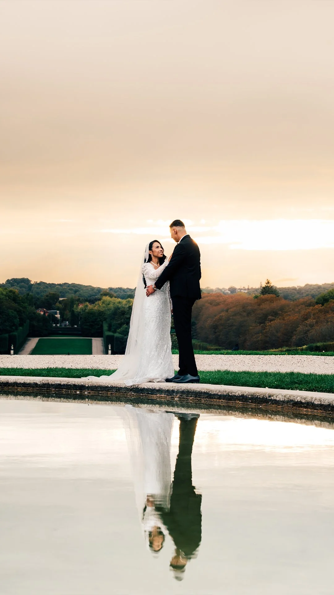 A bride in a white wedding dress and a groom in a black tuxedo holding hands, standing on a stone ledge near a reflecting pool during sunset. There are trees and a distant building in the background.