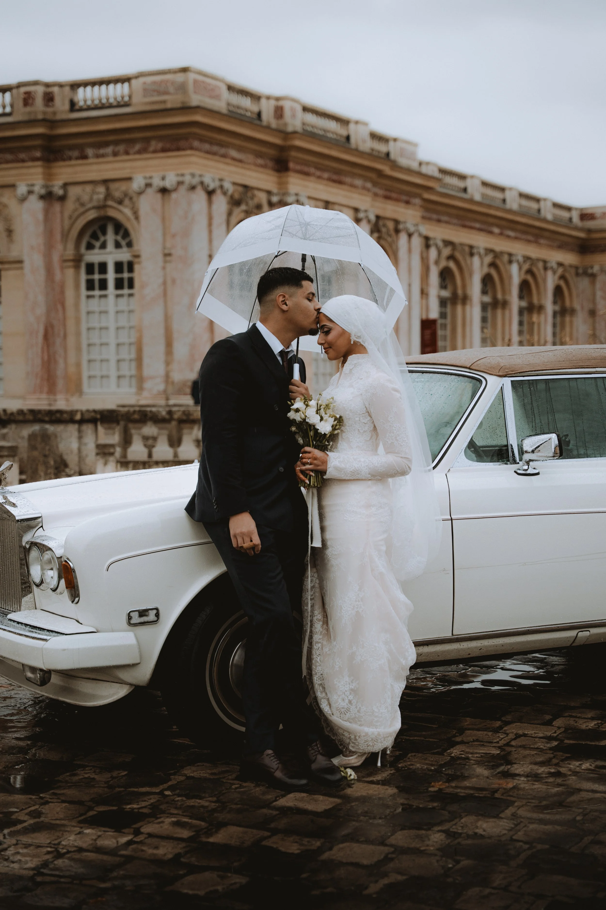 A bride and groom sharing a romantic moment under a transparent umbrella in front of a vintage white convertible car, with a historic building in the background on a rainy day.
