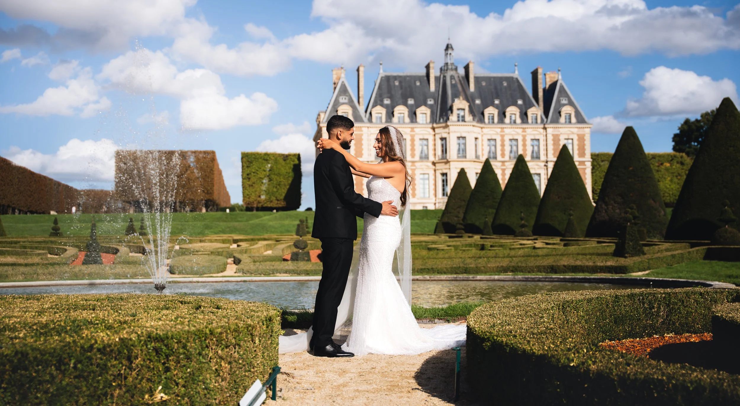 A bride and groom standing close together in front of a castle-like building, smiling and holding each other, during a wedding photoshoot outdoors on a sunny day with a fountain and manicured gardens in the background.