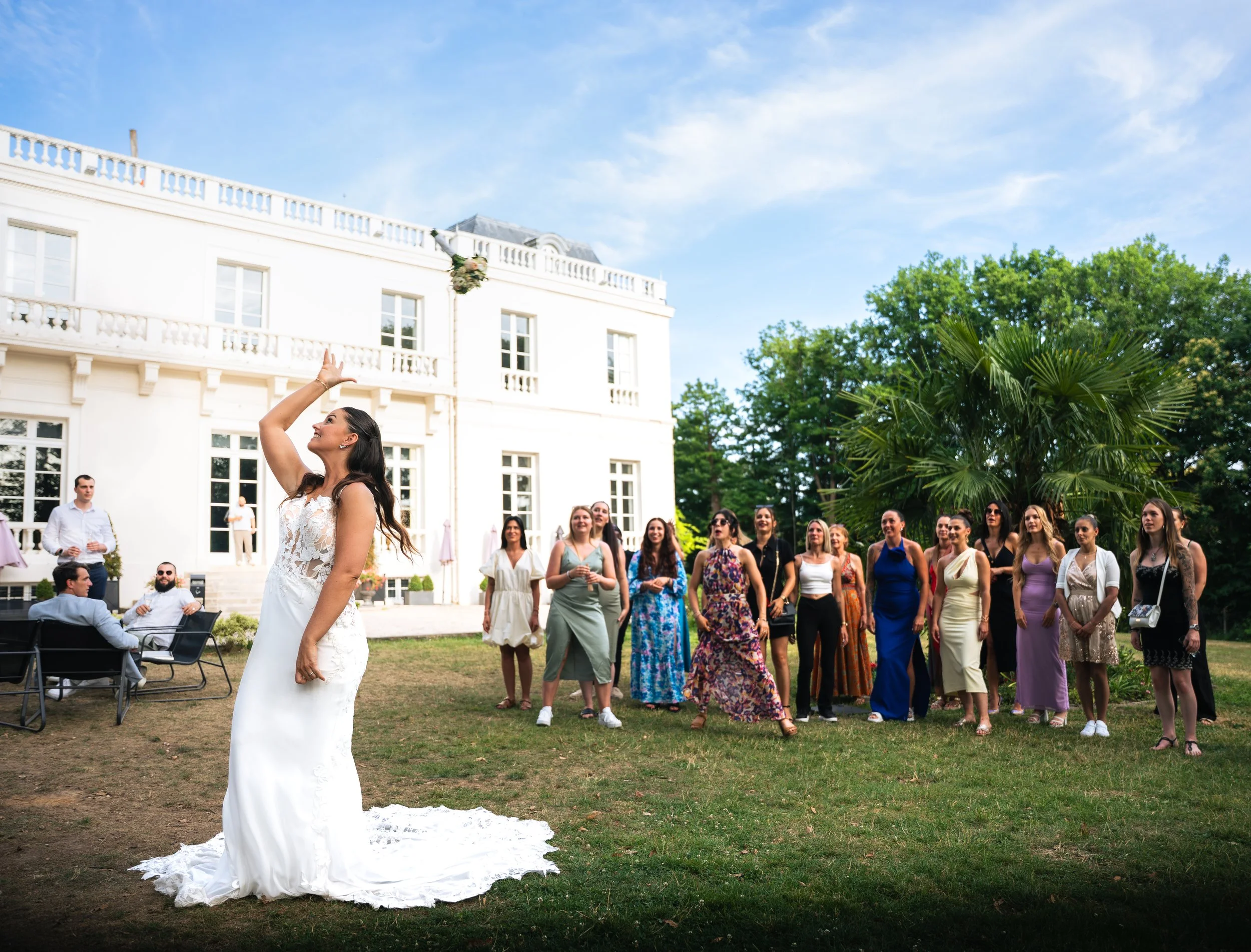 A bride in a white wedding dress throws her bouquet into the air during a wedding celebration outdoors, while a group of women and men look on happily.