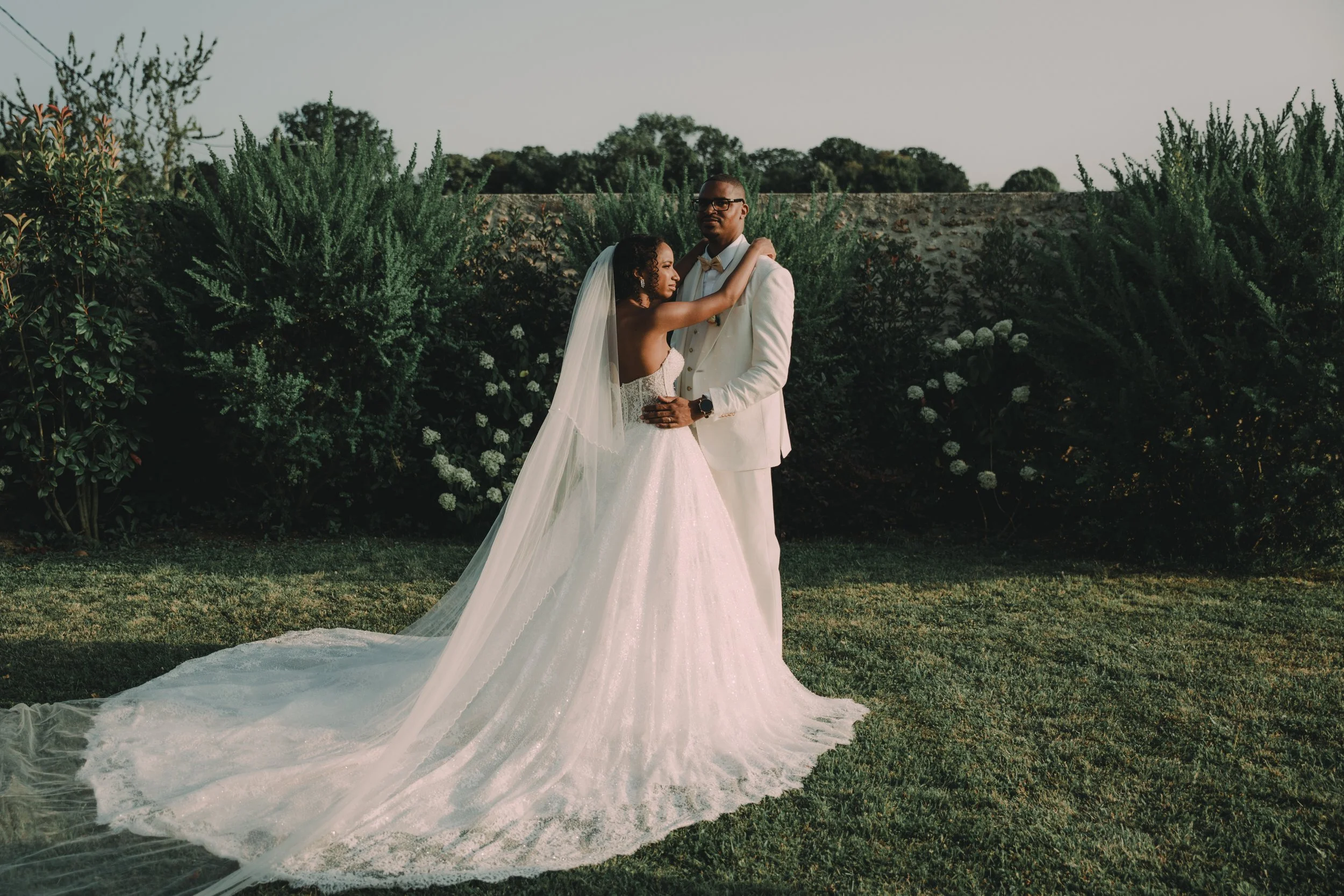 A bride and groom embracing outdoors, with a backdrop of green bushes and trees, during sunset.