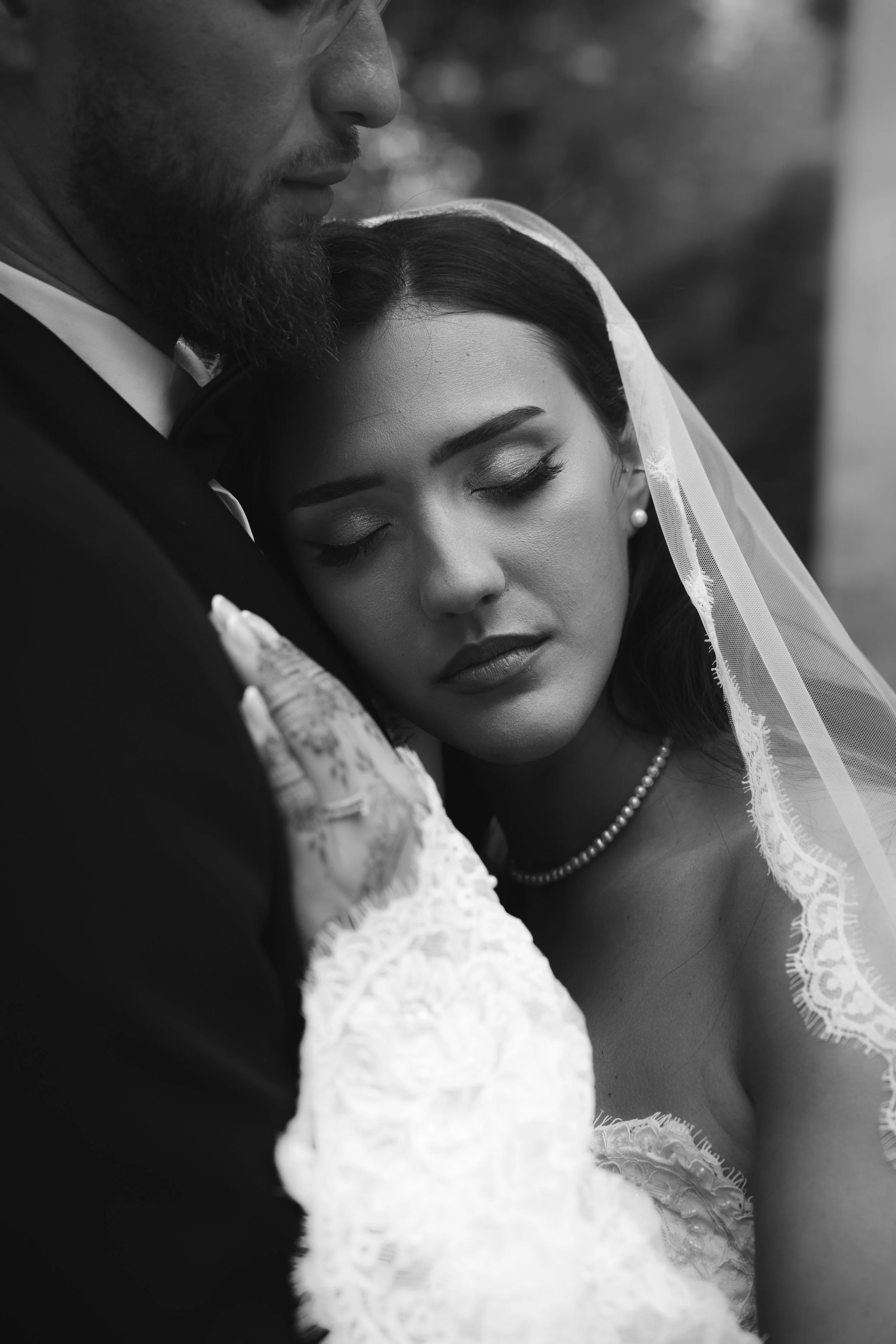 Un homme et une femme en robe de mariage, la femme avec un voile et des perles, semblent partager un moment intime et émotionnel lors d'une cérémonie ou d'une séance photo, en noir et blanc.
