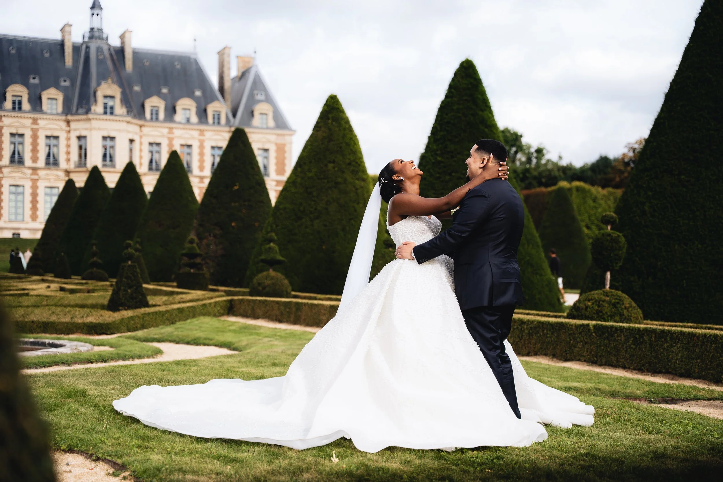 A bride and groom dancing outdoors in front of a castle with tall green trees.